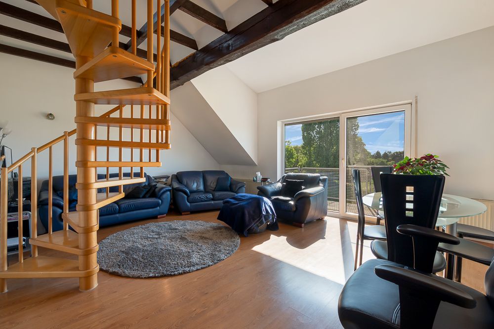 Wohnzimmer mit Holz-Wendeltreppe, blauen Sofas, Teppich und großen Fenstern mit Blick nach draußen.