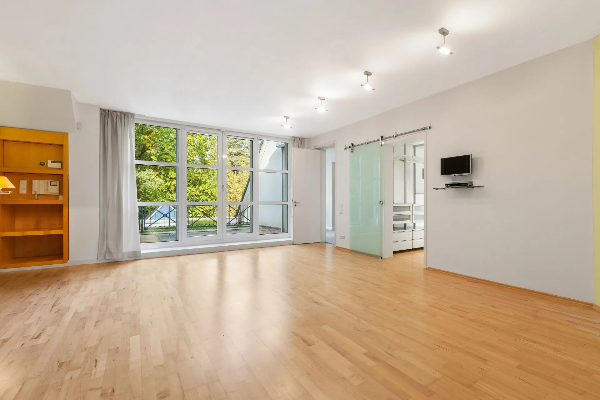 Helles Wohnzimmer mit Holzboden, großen Fenstern, Regalfach und Glastür.