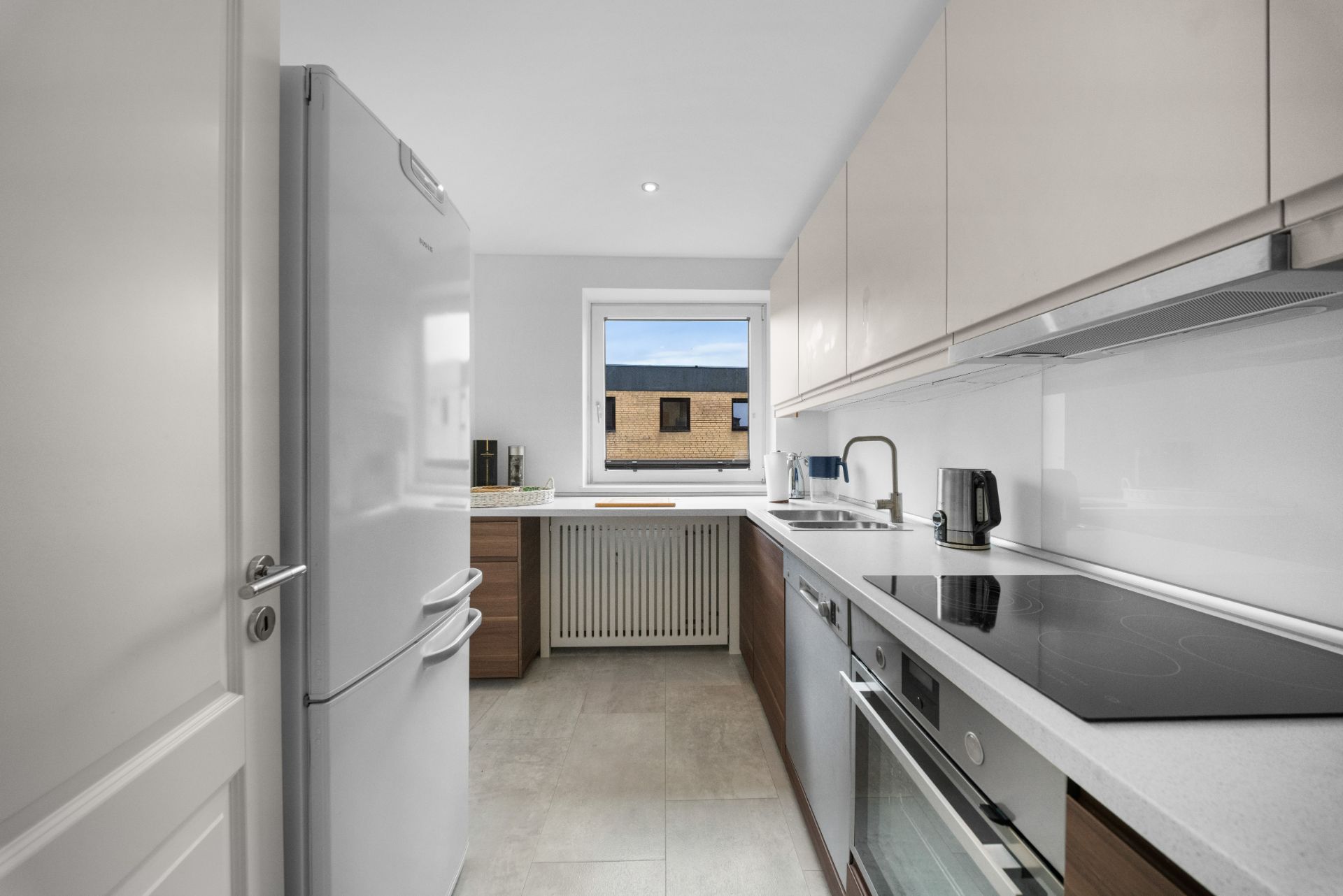 Moderne, schmale Küche mit weißen Schränken, Elektrogeräten und Fenster am Ende des Raumes.