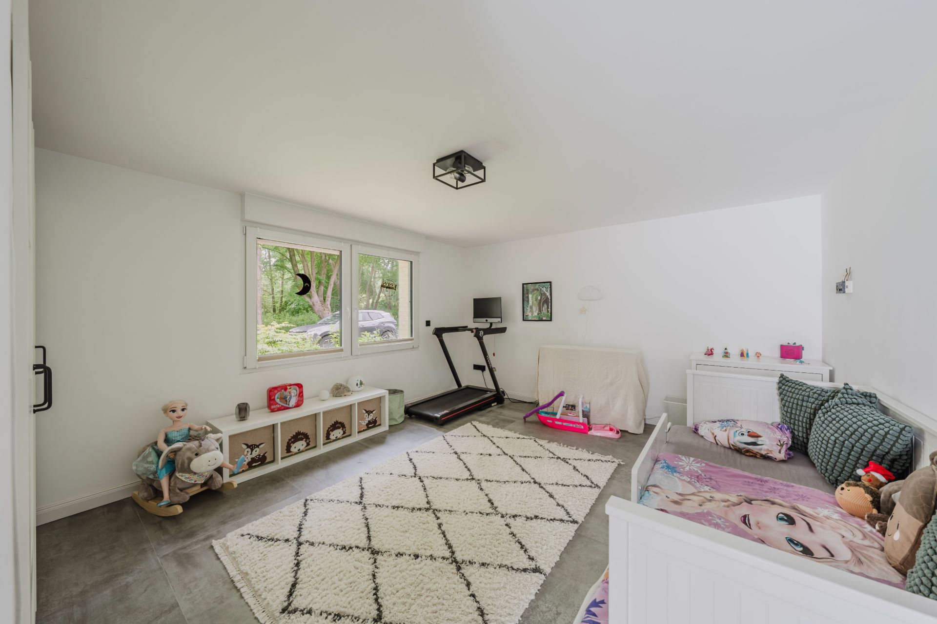 Helles Kinderzimmer mit Teppich, Spielzeug, Laufband und Tagesbett, Fenster mit Blick ins Grüne.