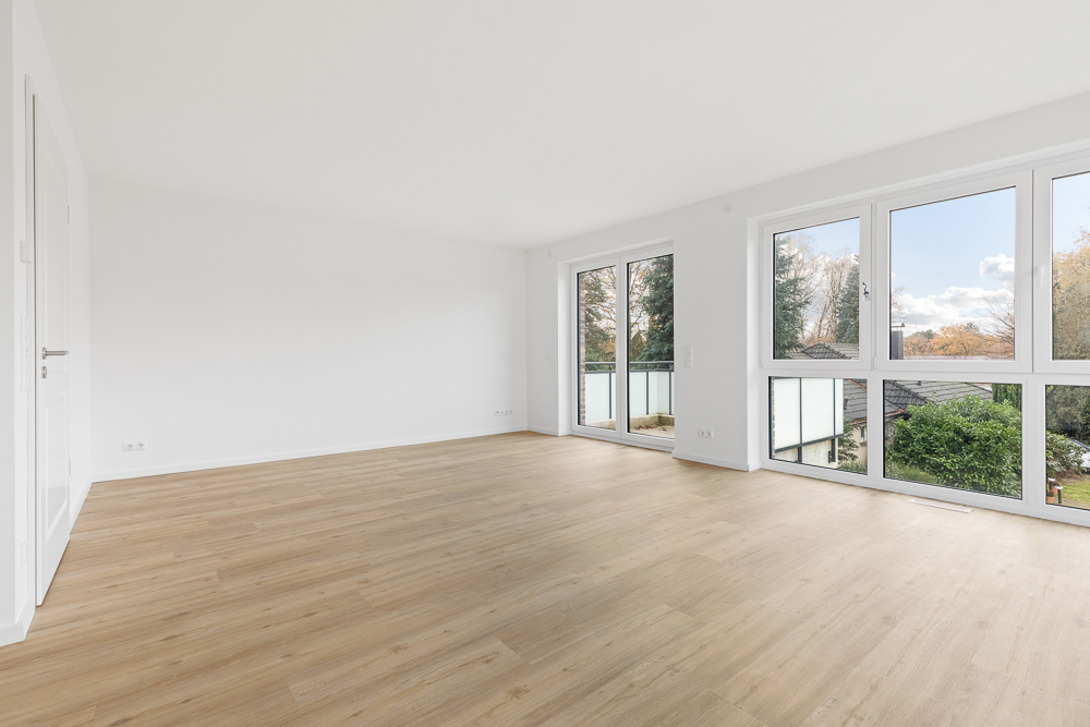 Heller, leerer Raum mit Holzfußboden und großen Fenstern mit Blick ins Grüne.