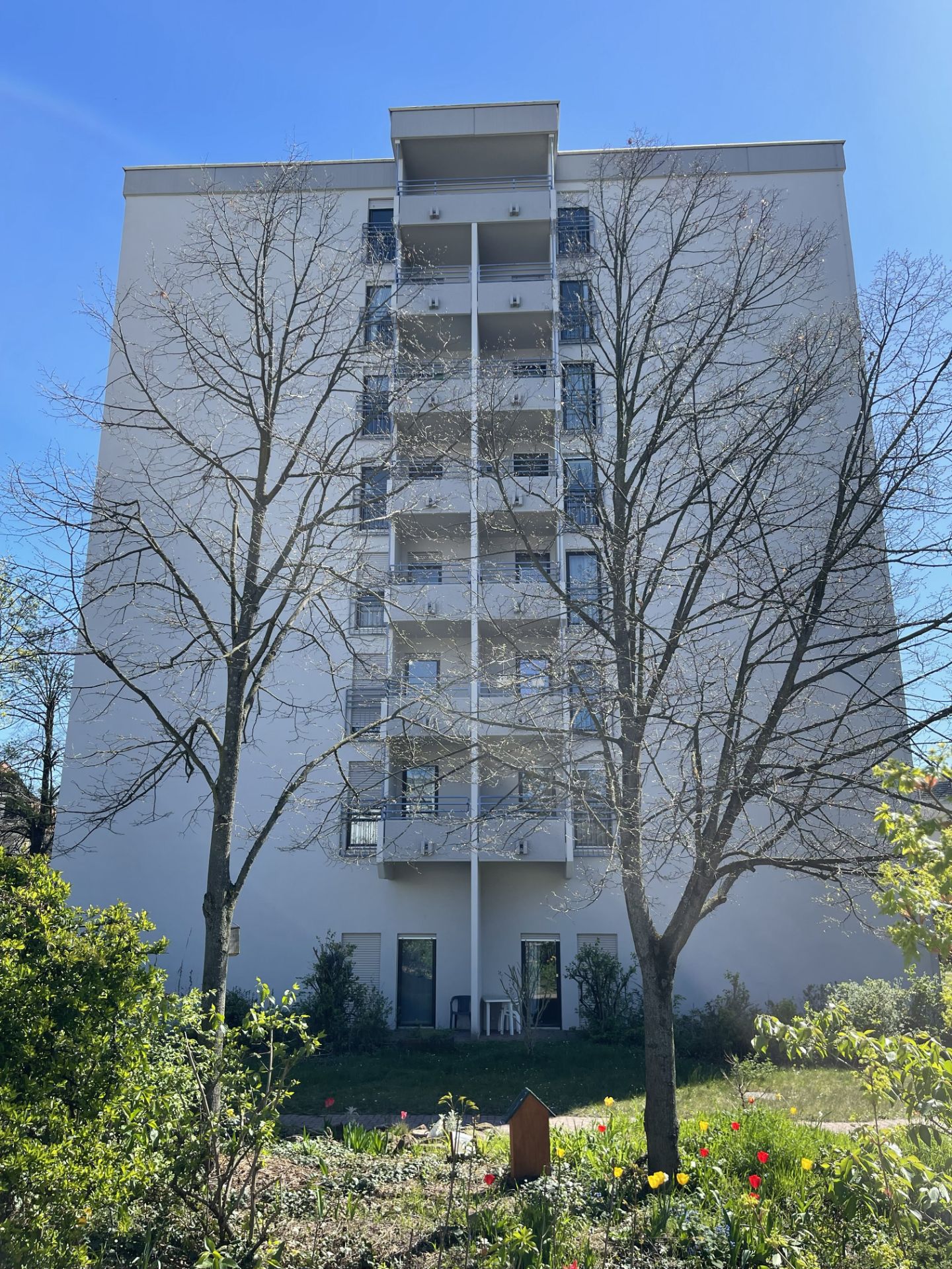 Mehrstöckiges Wohnhaus mit Balkonen und Garten im Vordergrund, Bäume ohne Blätter.