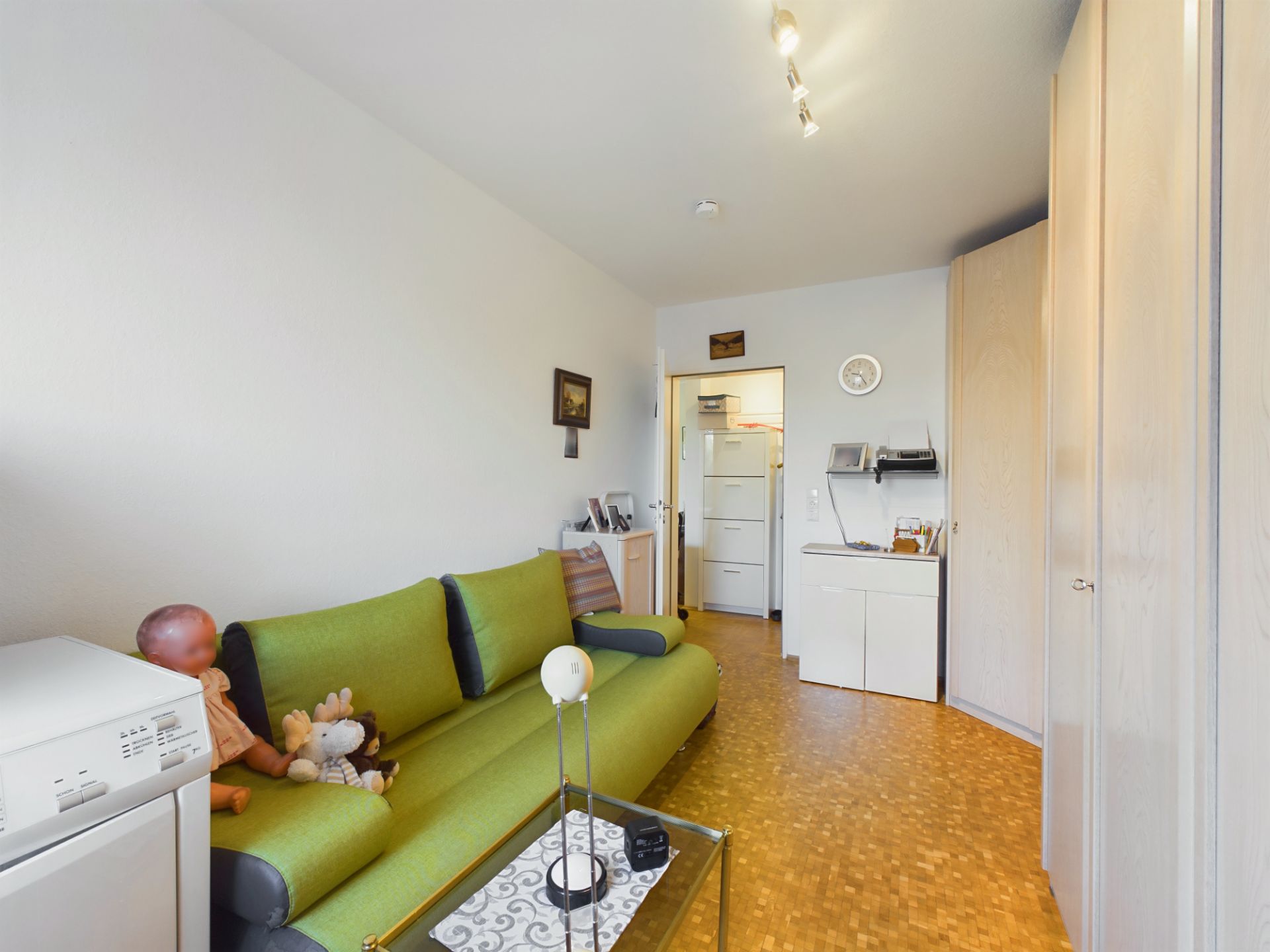 Wohnzimmer mit grünem Sofa, Glastisch, Schrankwand und hellen Holzmöbeln.