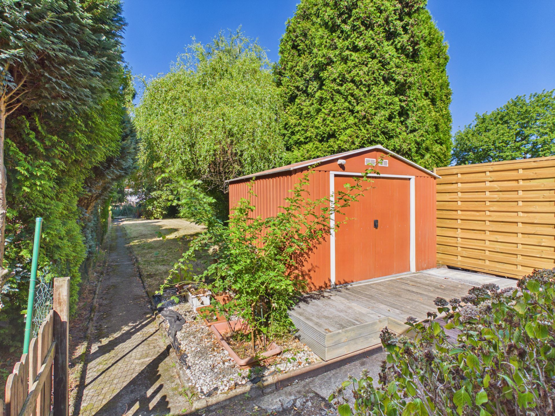 Rotes Gartenhaus auf Holzterrasse, umgeben von Bäumen und Holzlattenzaun.