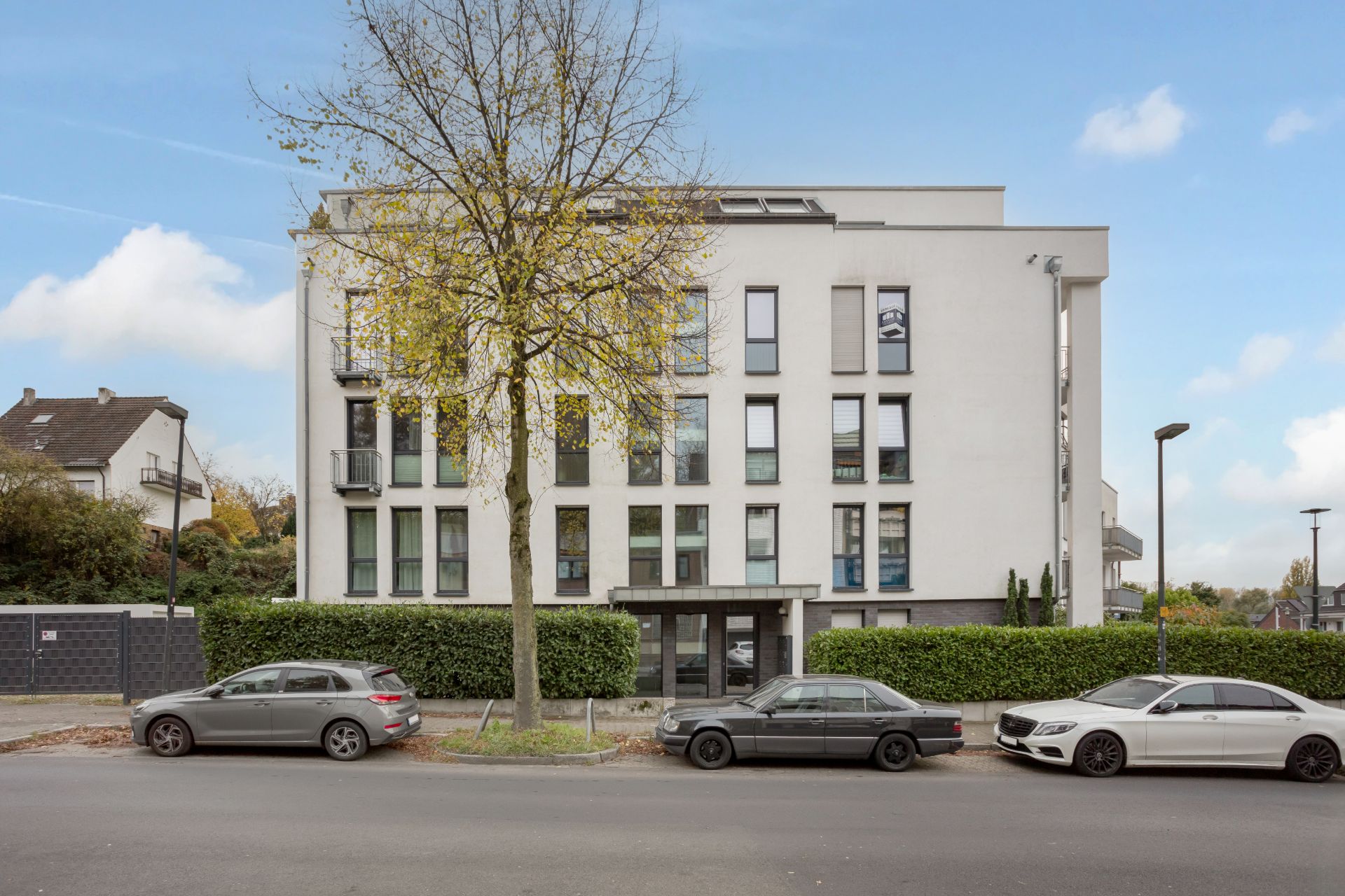 Modernes, weißes Wohnhaus mit mehreren Etagen, davor drei parkende Autos und ein Baum.