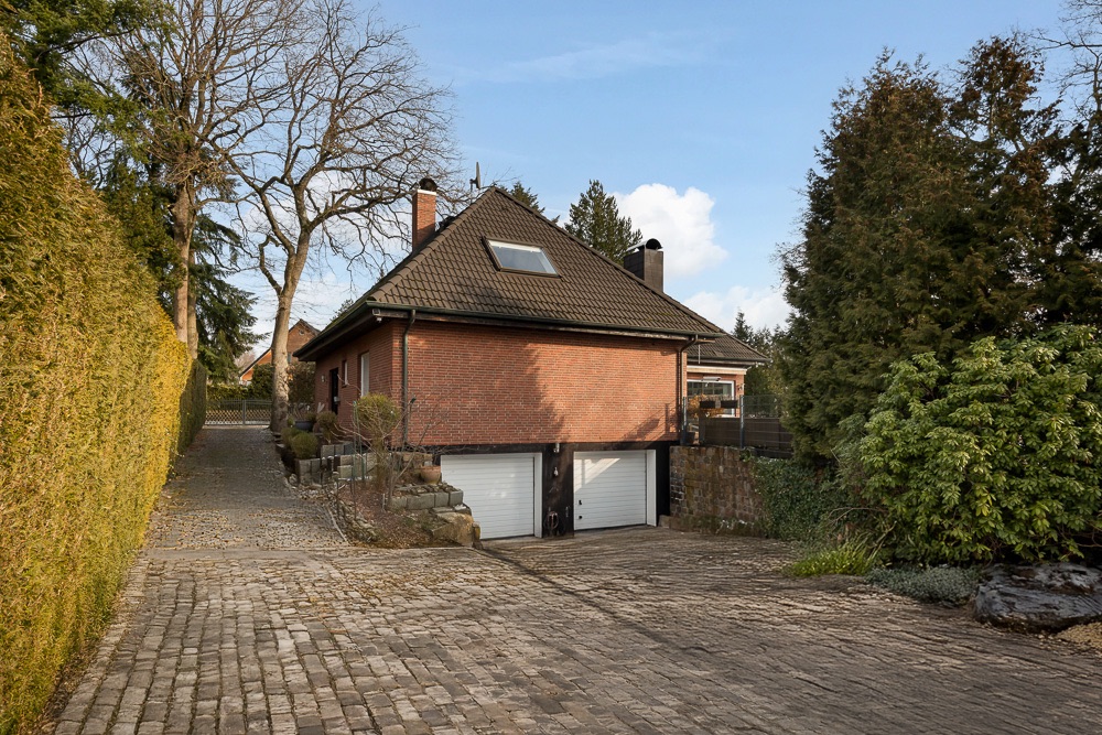Backsteinhaus mit dunklem Dach, großer Einfahrt und zwei Garagentoren, umgeben von Bäumen.