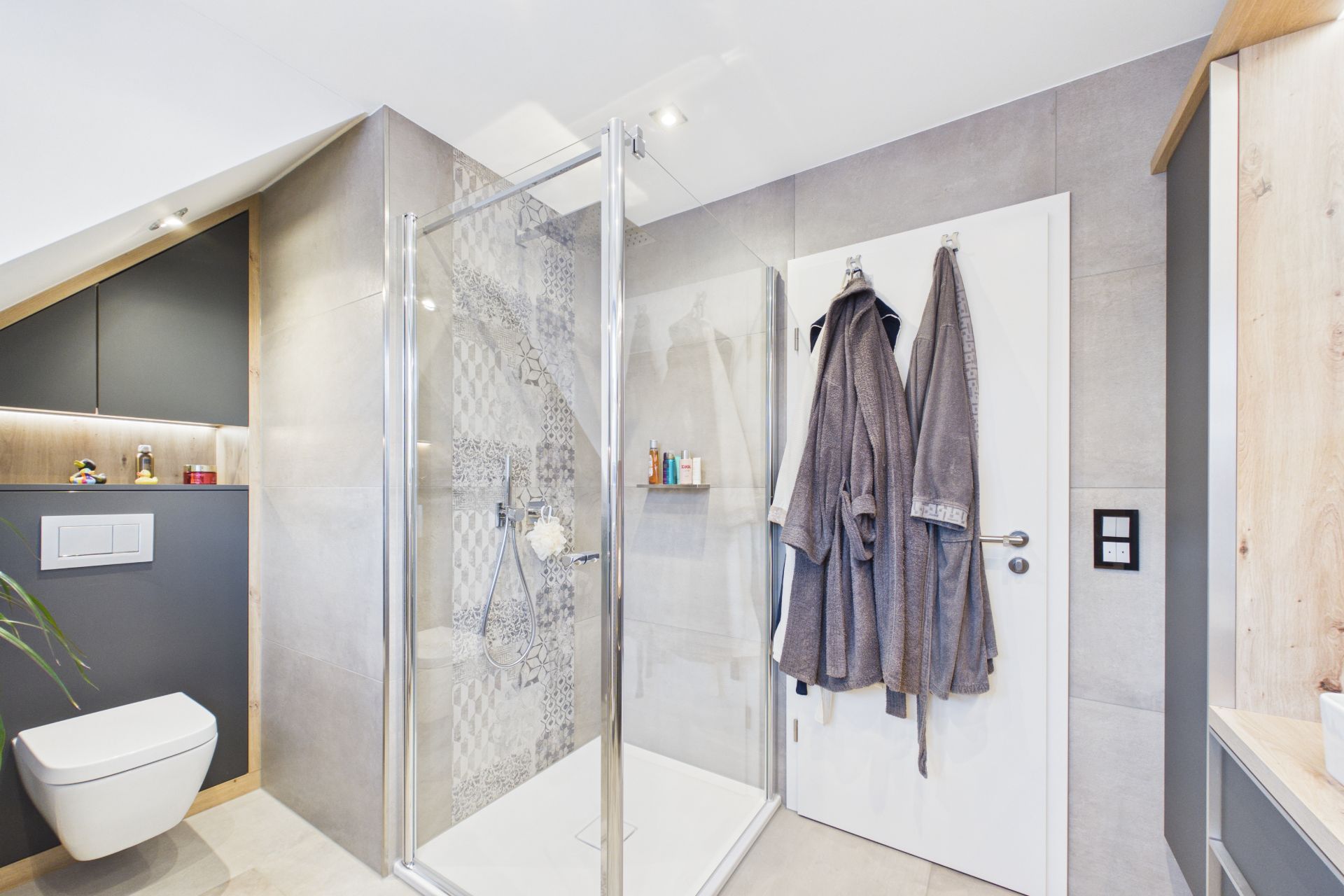 Modernes Badezimmer mit Glasdusche, WC, grauen Fliesen und zwei Bademänteln an der Tür.