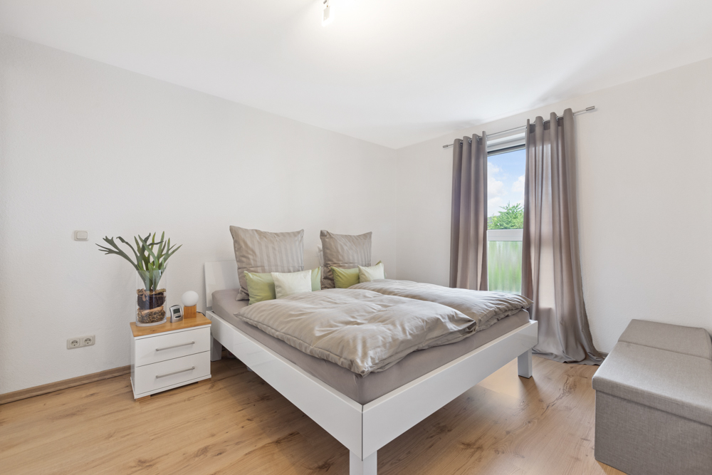 Helles Schlafzimmer mit Doppelbett, Nachttisch, Pflanze und Fenster mit grauen Vorhängen.