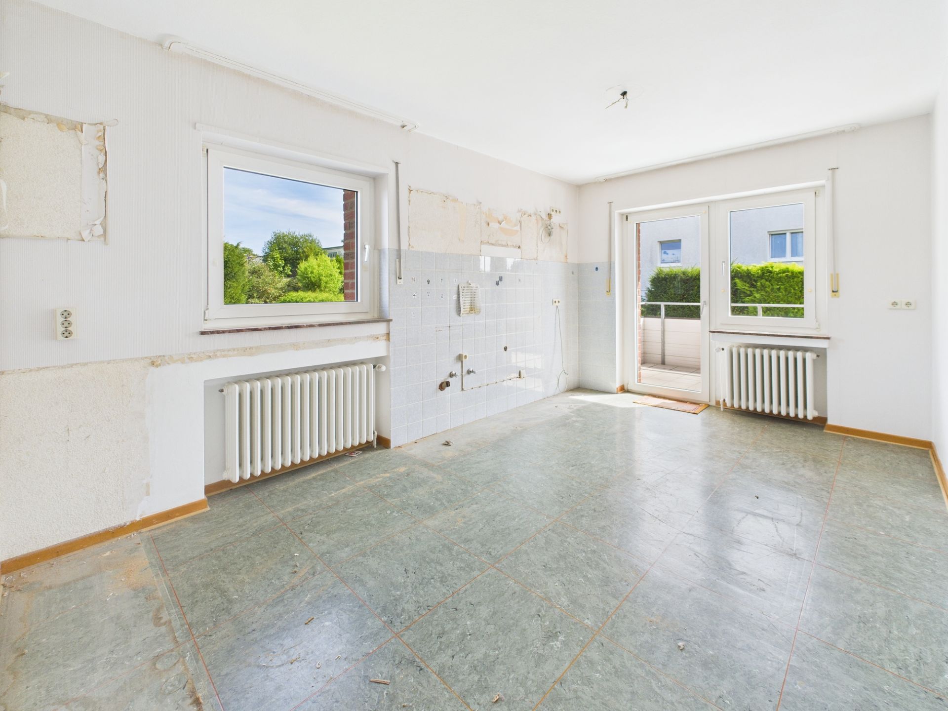 Leerer Raum mit zwei Fenstern, einer Tür zum Garten und zwei Heizkörpern.