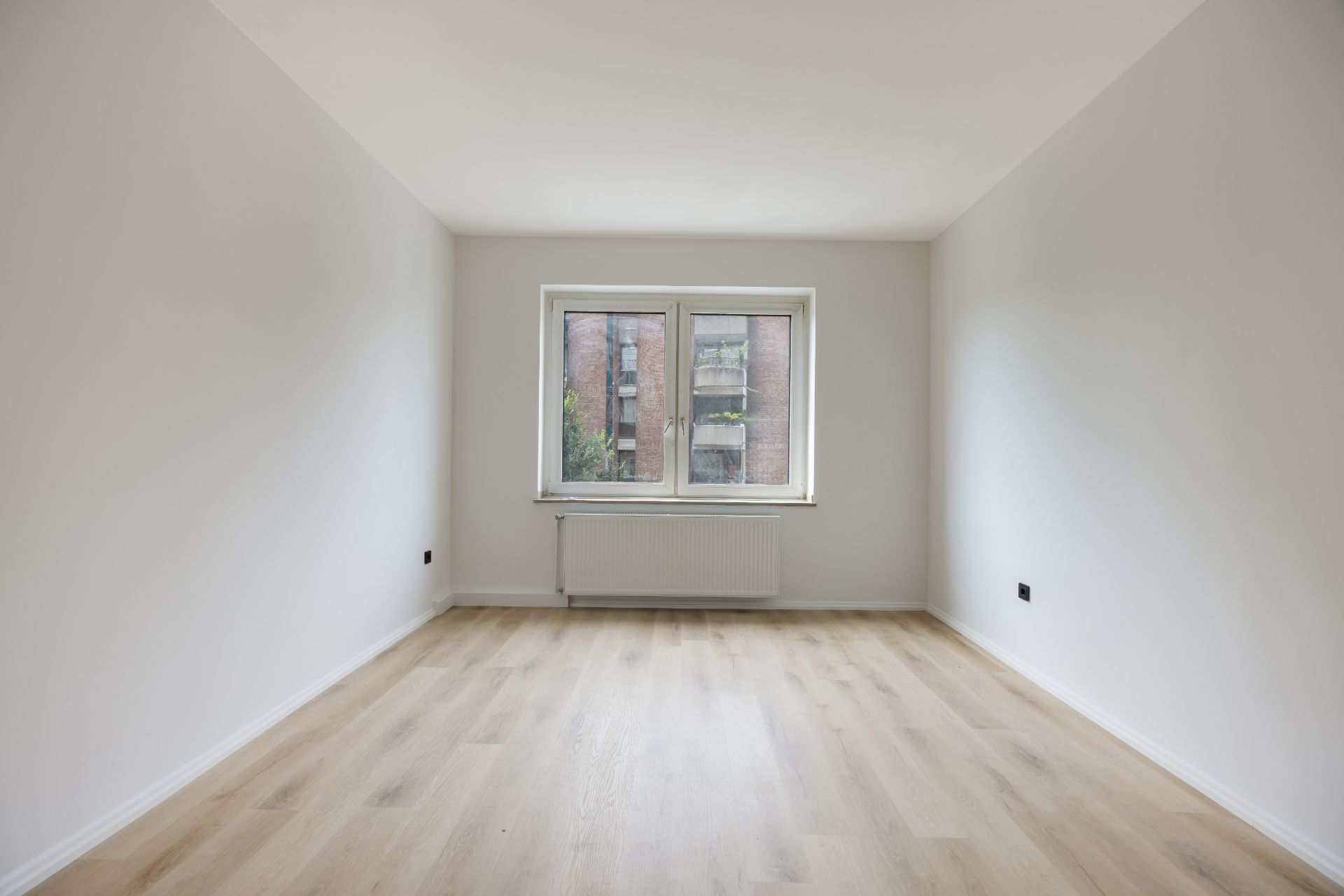 Leerer Raum mit hellem Holzboden, weiß gestrichenen Wänden und einem Fenster mit Blick nach draußen.