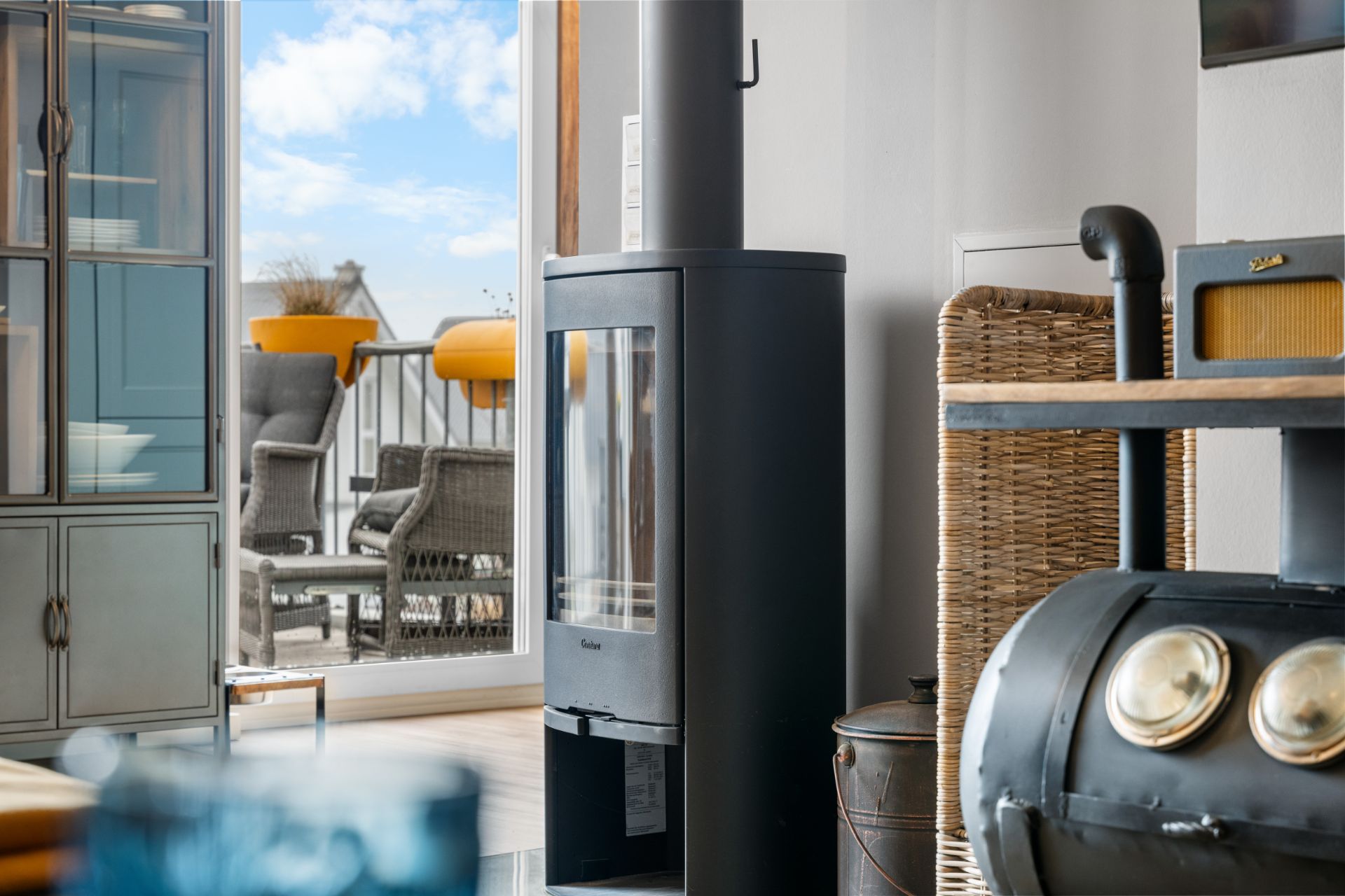 Ein moderner Kaminofen im Wohnzimmer mit Blick auf eine Terrasse mit Stühlen und Pflanzkübeln.