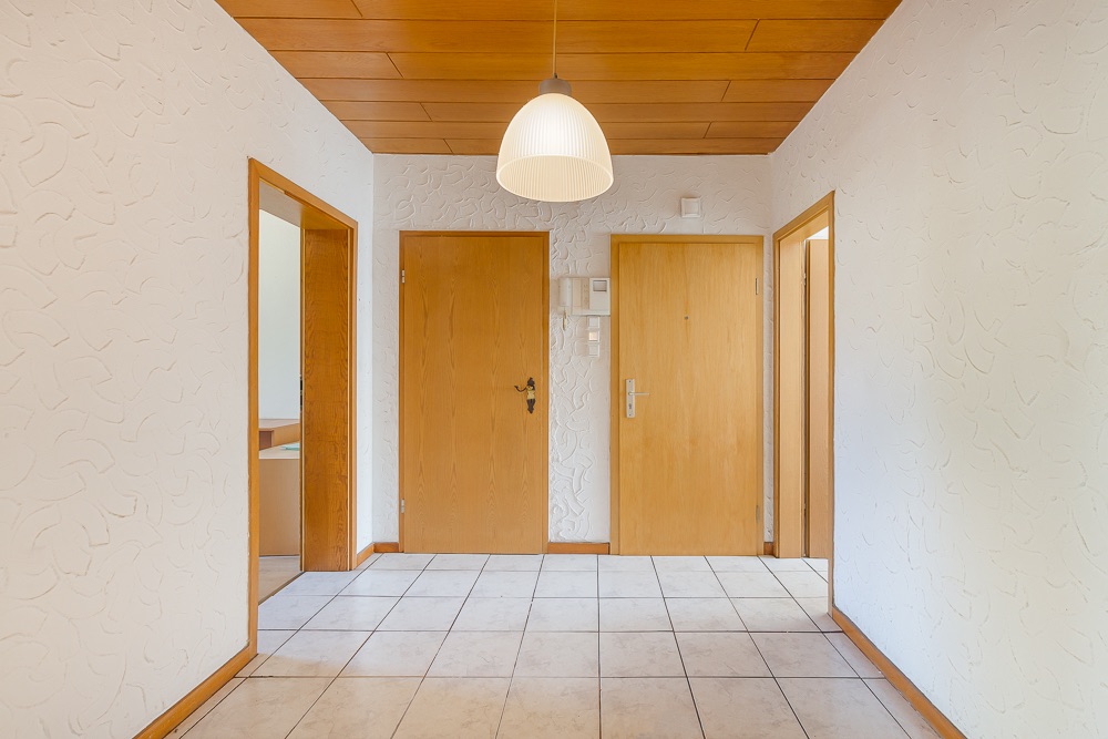 Heller Flur mit Fliesenboden, Holzdecke, drei Türen und einer Deckenlampe.