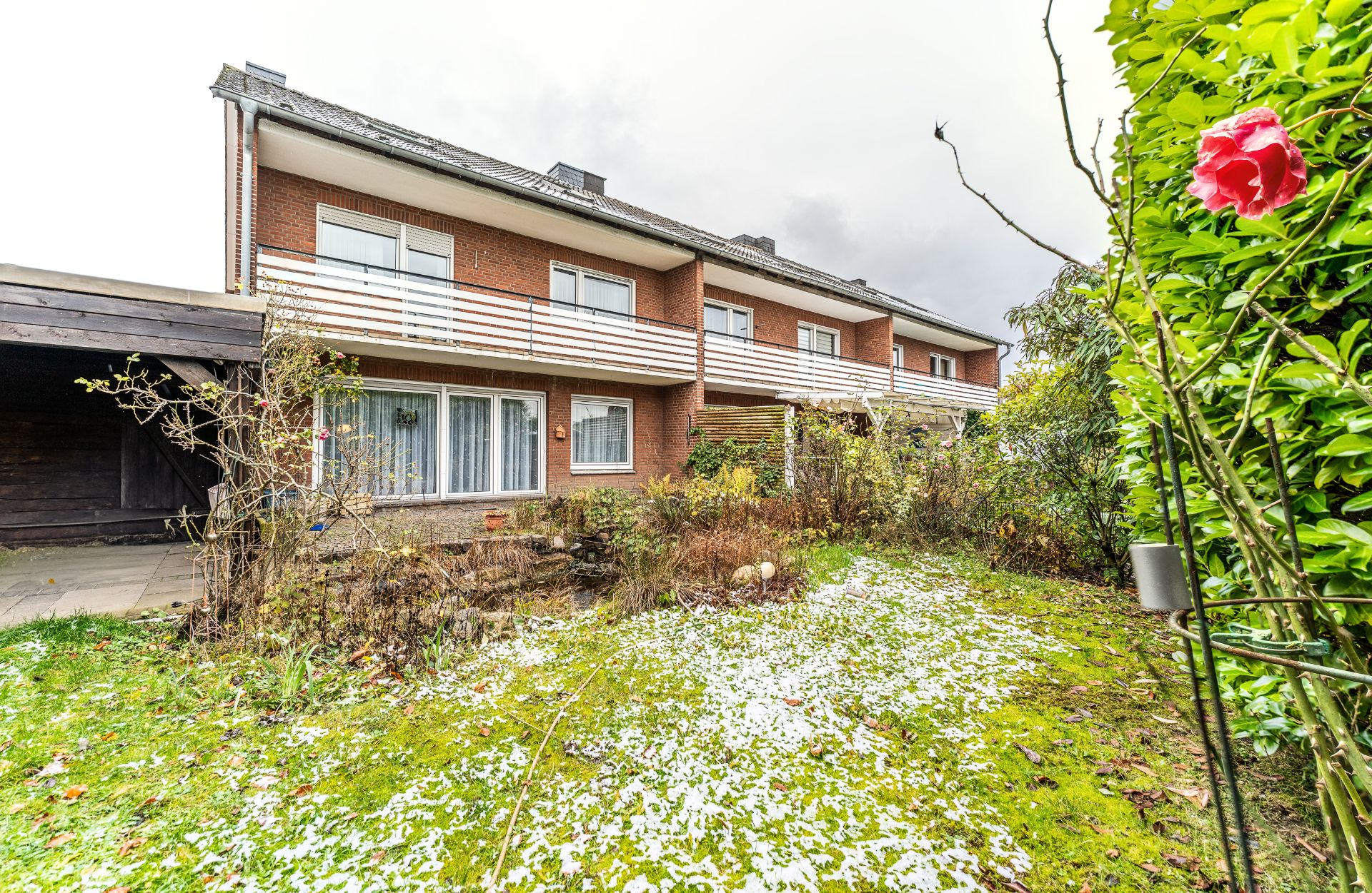 Reihenhaus mit Balkon und kleinem Garten, teils mit Schnee bedeckt, viele Sträucher und eine Rose.