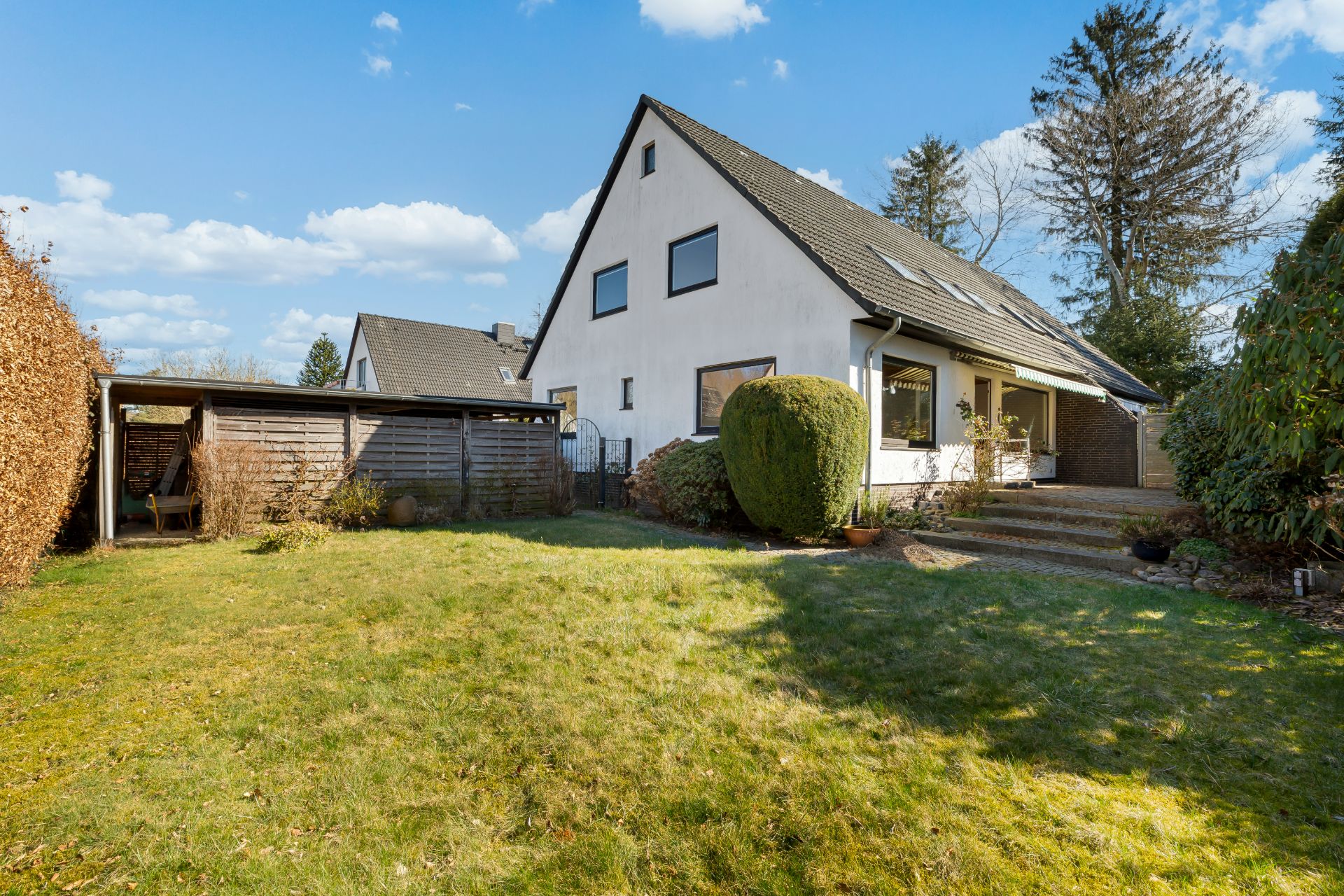 Einfamilienhaus mit Satteldach, Garten und Carport, umgeben von Sträuchern und Bäumen.