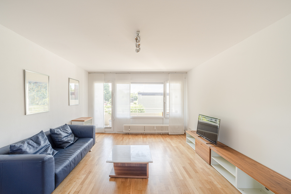 Helles Wohnzimmer mit blauem Sofa, Couchtisch, TV und großen Fenstern mit Vorhängen.