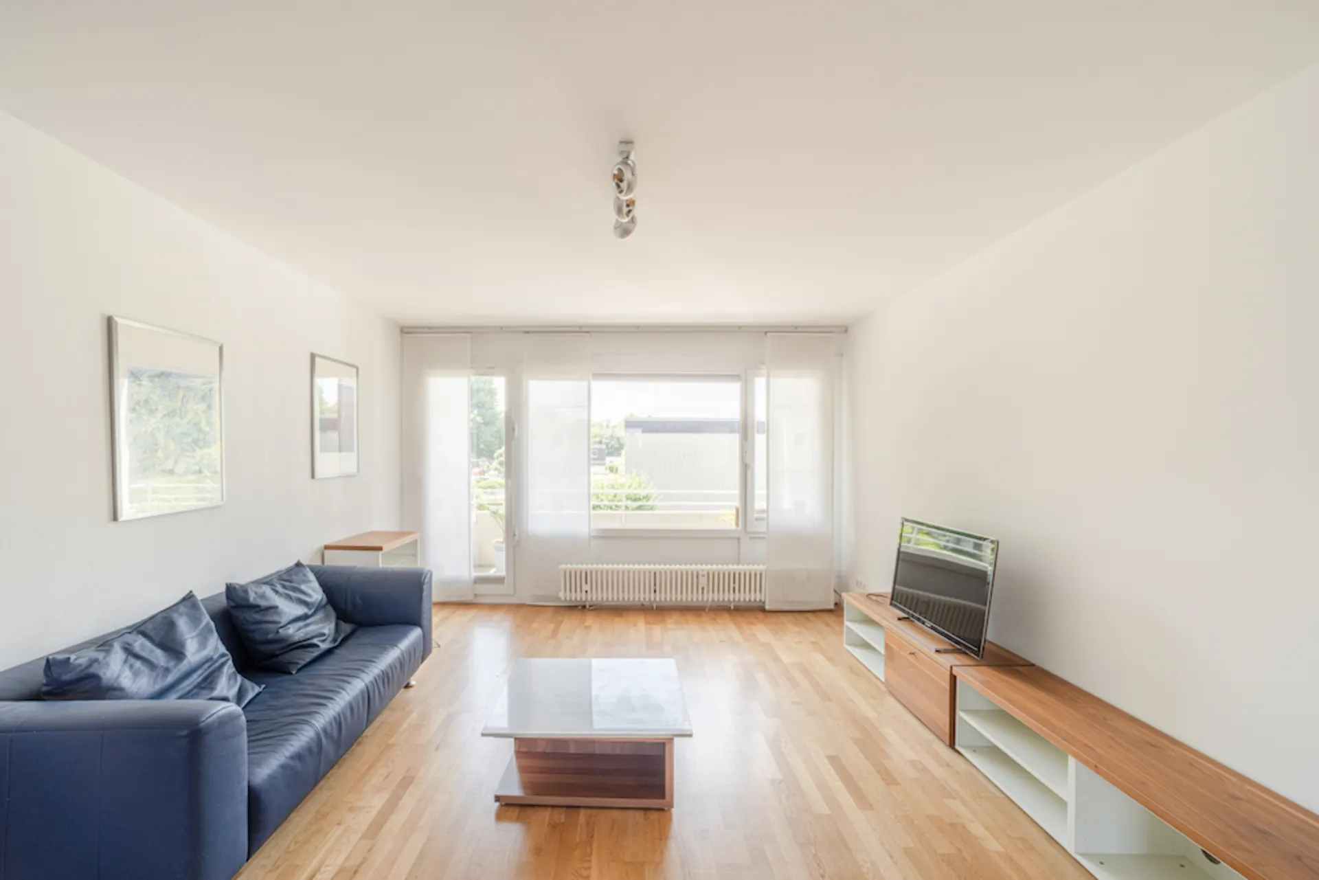 Helles Wohnzimmer mit blauem Sofa, Couchtisch, TV und großen Fenstern mit Vorhängen.