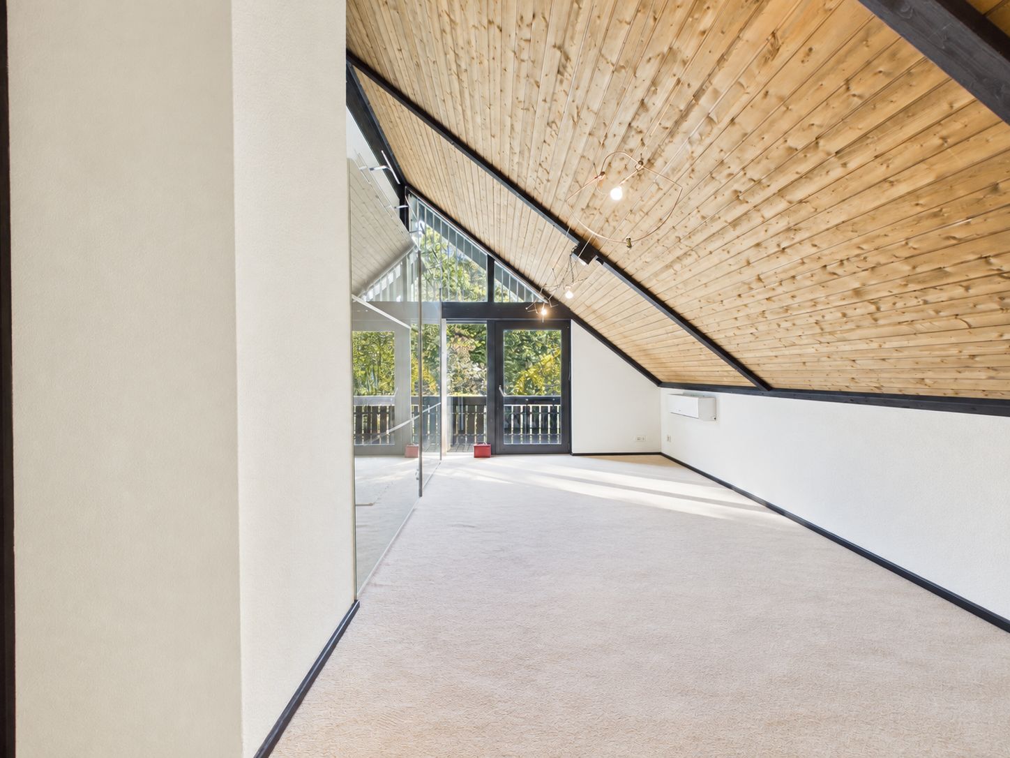 Heller Dachbodenraum mit schräger Holzdecke, Teppichboden und großen Fenstern zum Balkon.