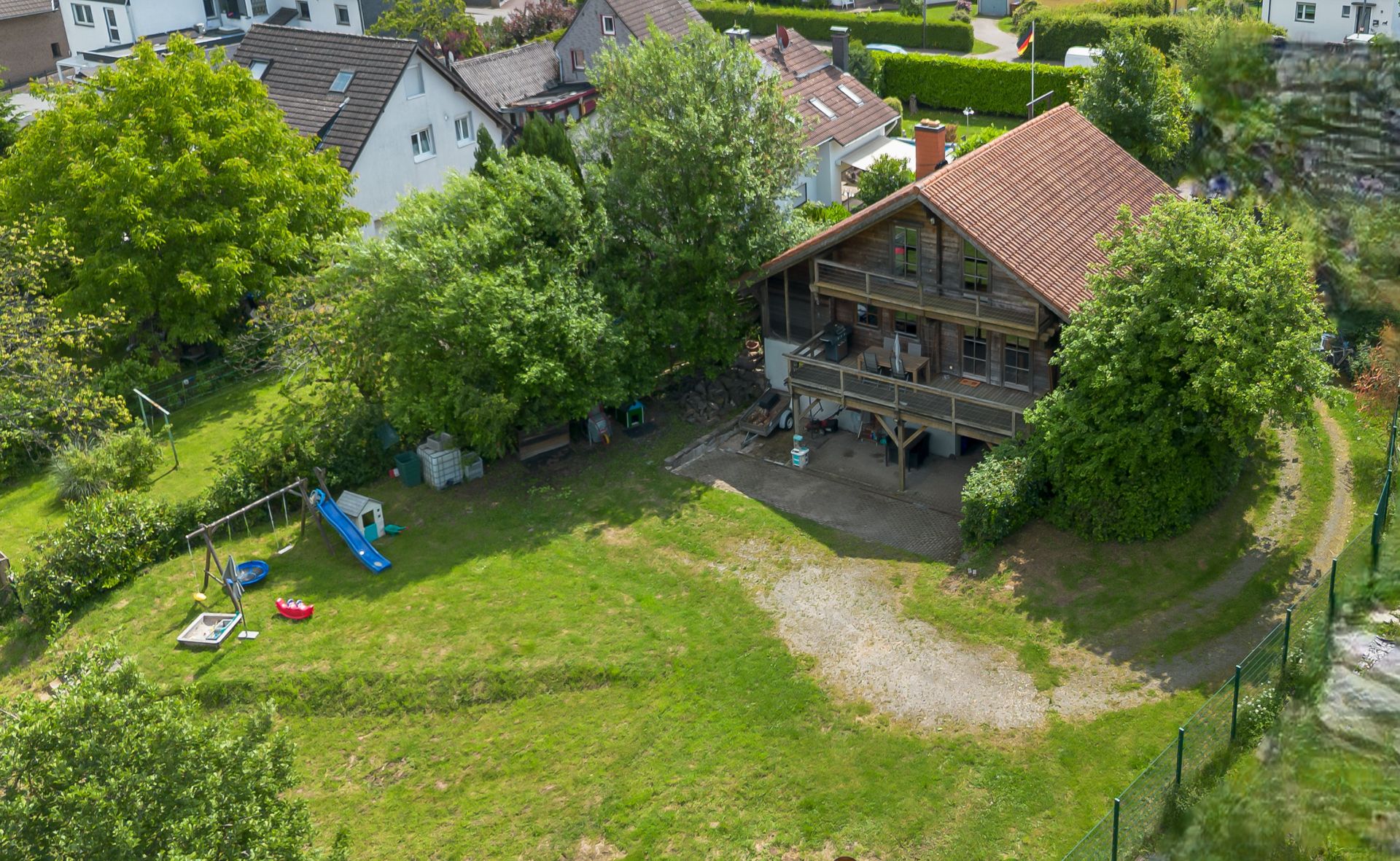 Holzhaus mit Balkon und großem Garten, Spielplatz mit Rutsche und Sandkasten, umgeben von Bäumen.