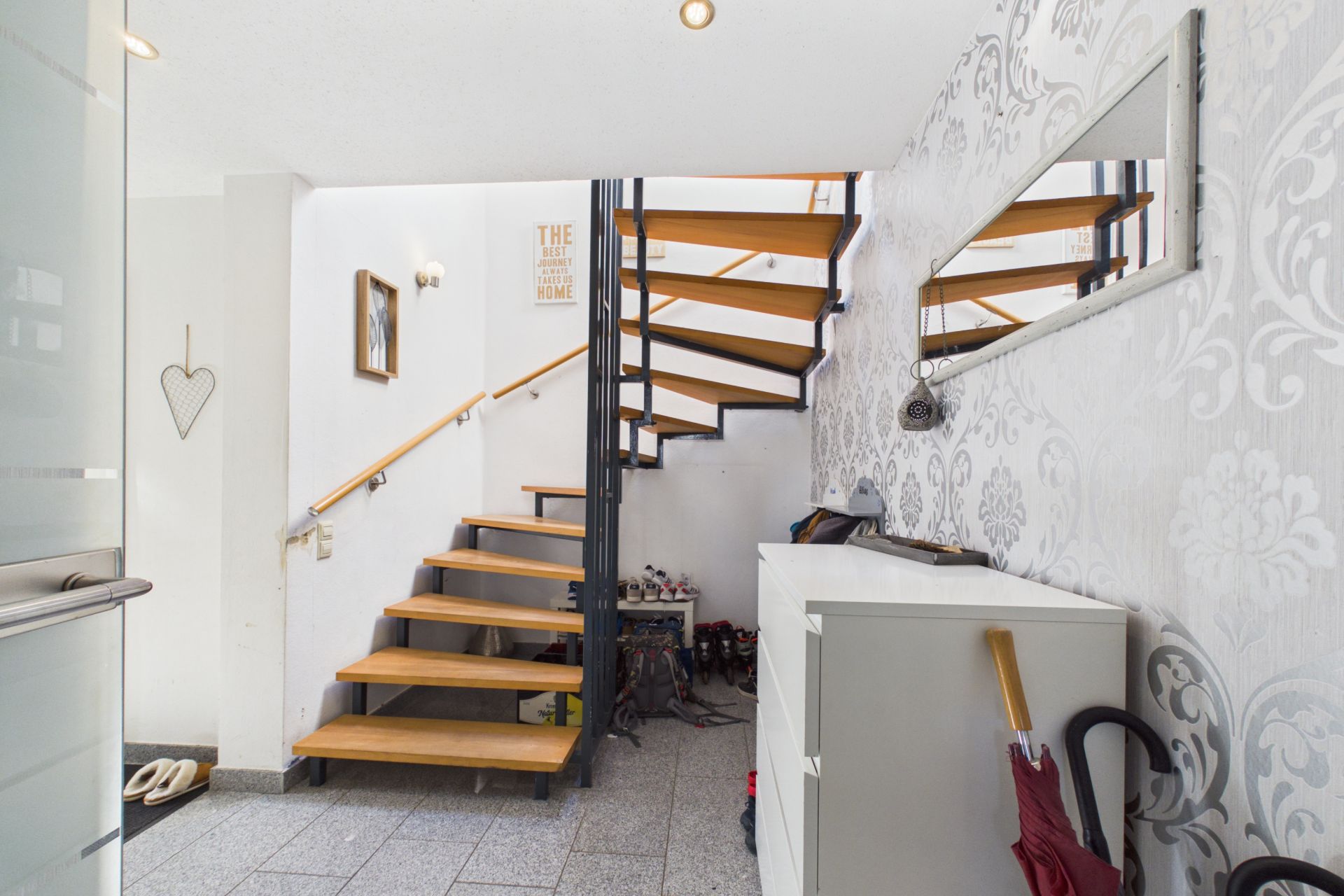 Flur mit offener Holztreppe, Kommode, Spiegel und Regenschirmen an der Wand.