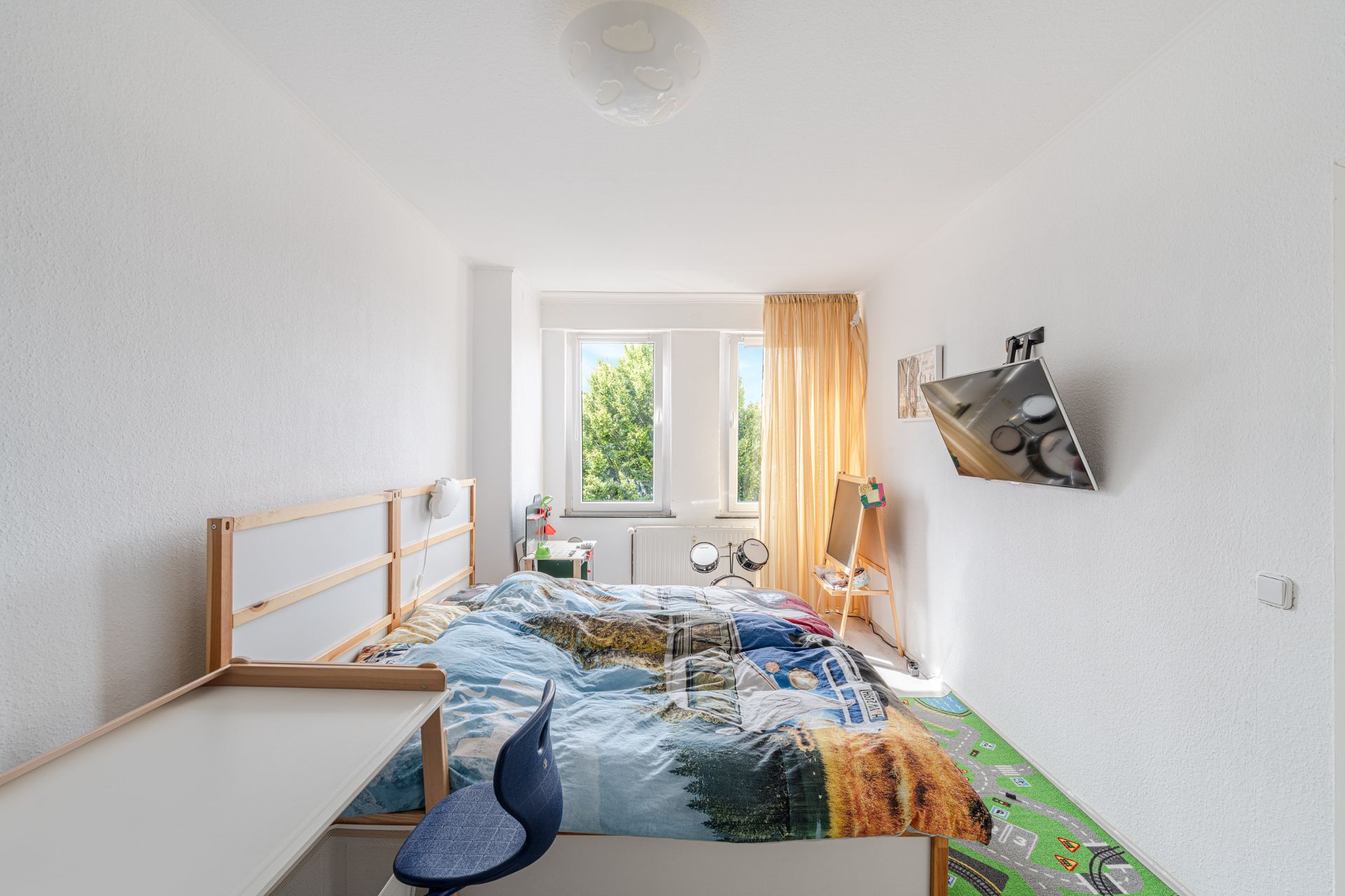 Helles Kinderzimmer mit Bett, Schreibtisch, Fernseher und zwei Fenstern mit gelben Vorhängen.