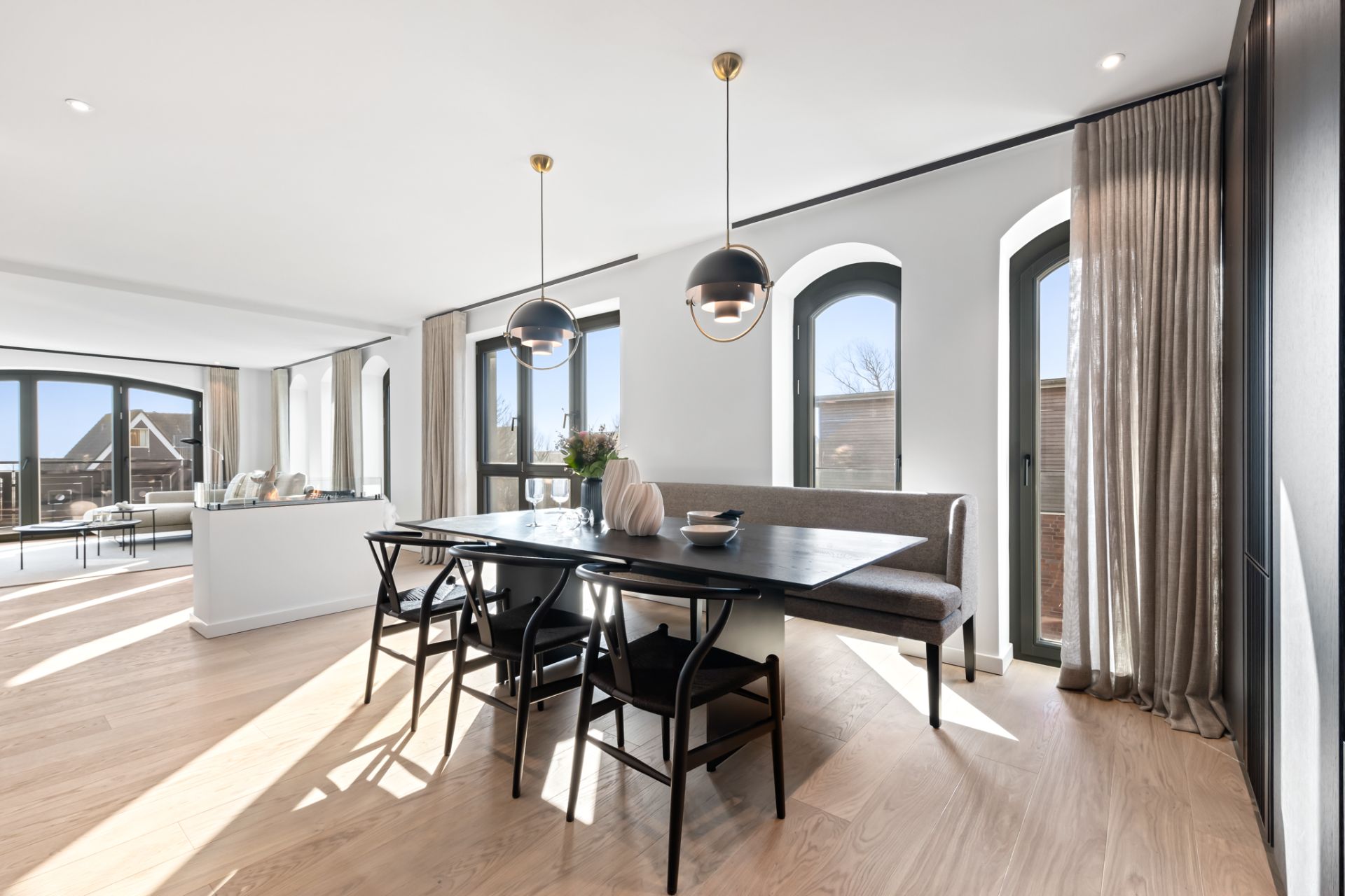 Helles Esszimmer mit schwarzem Tisch, Stühlen, Bank und zwei Hängeleuchten, große Fenster.