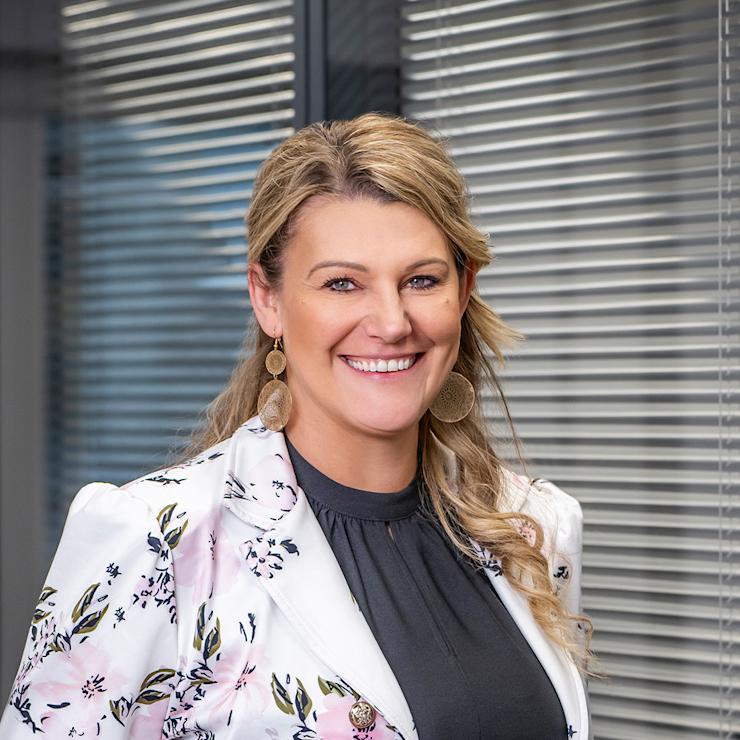 Frau mit blondem Haar, schwarzem Oberteil und weißem Blazer mit Blumenmuster, vor Jalousien stehend.