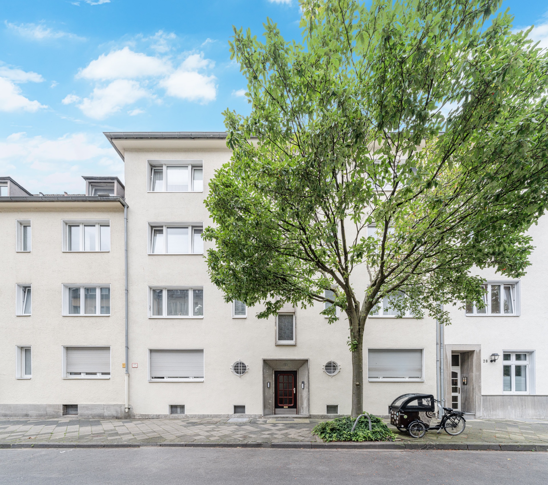 Mehrfamilienhaus mit hellen Fassaden, großem Baum davor und Lastenfahrrad auf dem Gehweg.