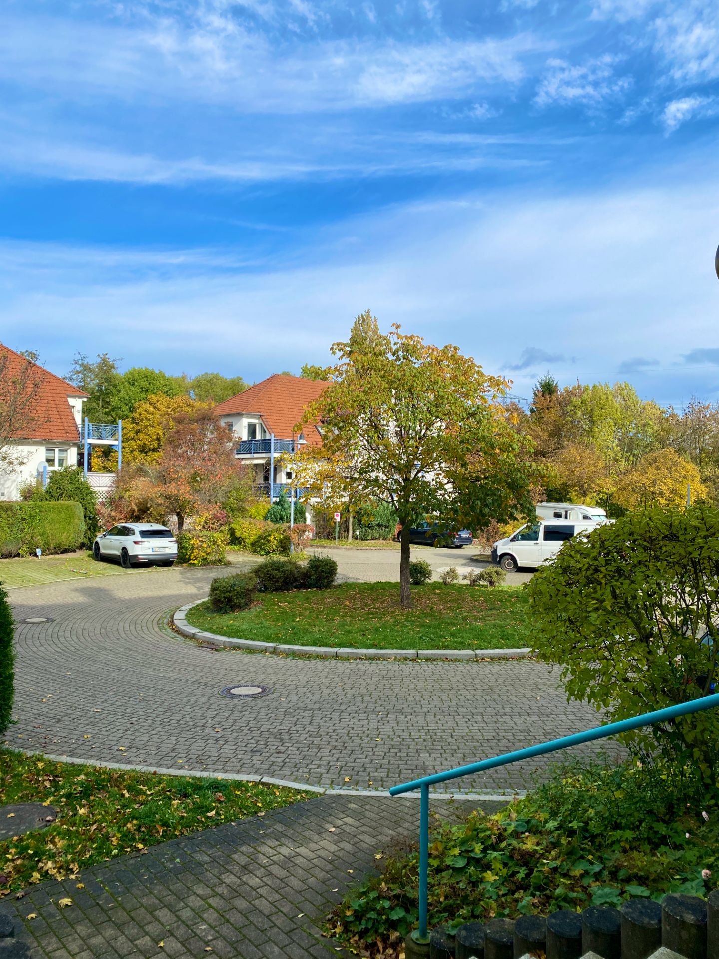Runder Wohnstraßenbereich mit Bäumen, geparkten Autos und Häusern mit roten Dächern bei blauem Himmel.