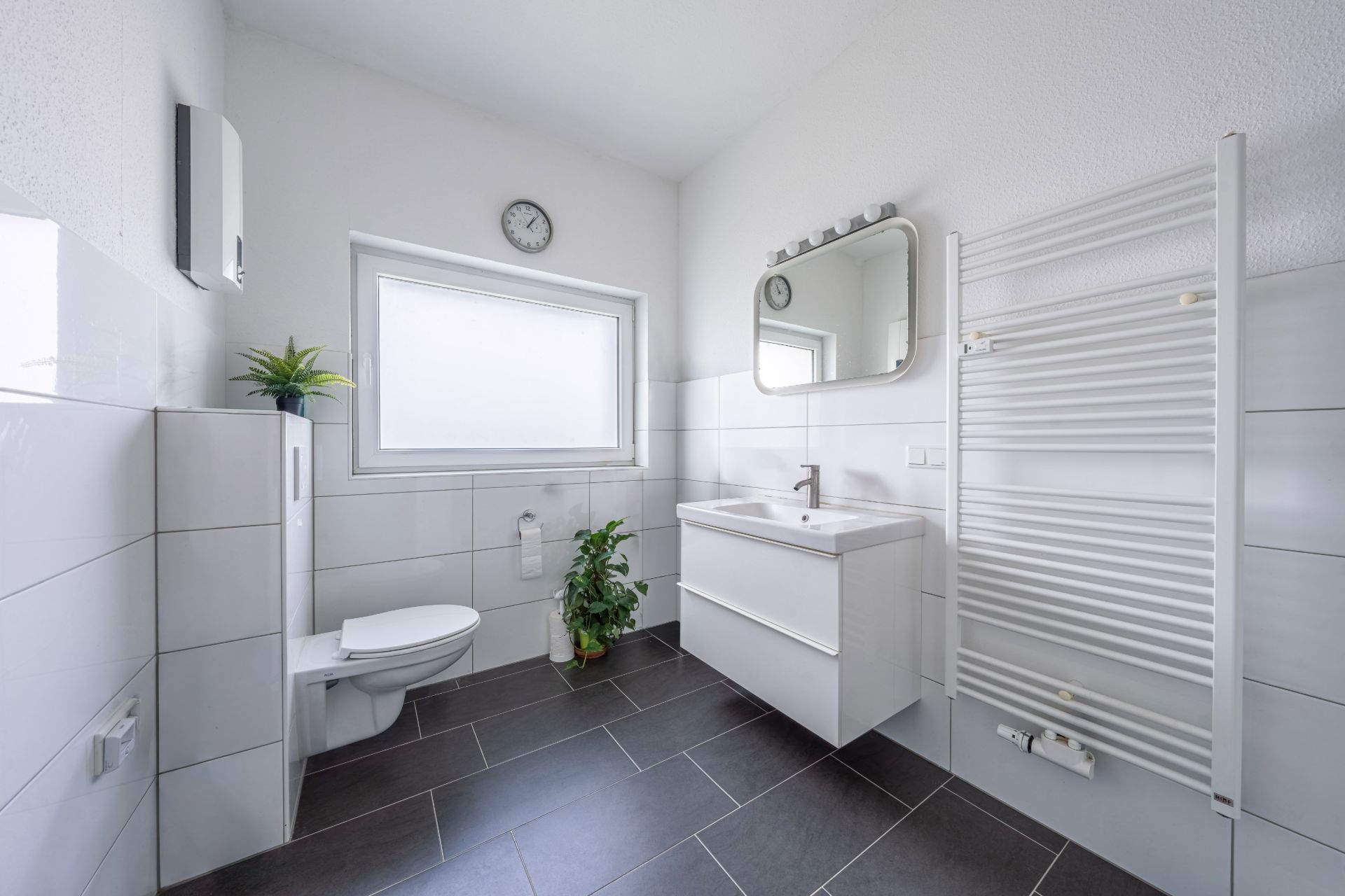 Modernes Bad mit WC, Waschbecken, Spiegel, Heizkörper, Pflanzen und großem Fenster.