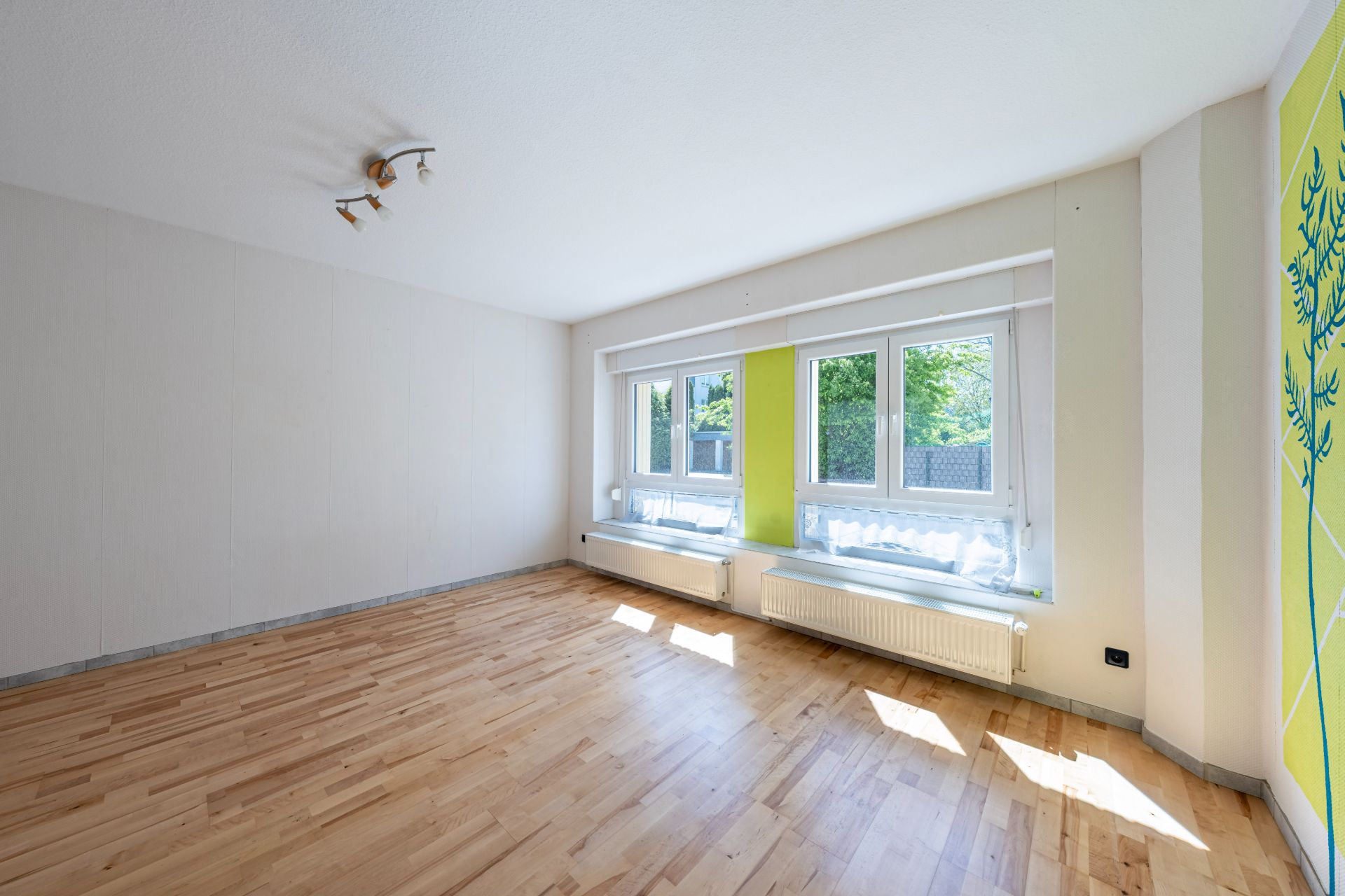Helles, leeres Zimmer mit Parkettboden, großen Fenstern und gelb-grün gestrichener Wand.