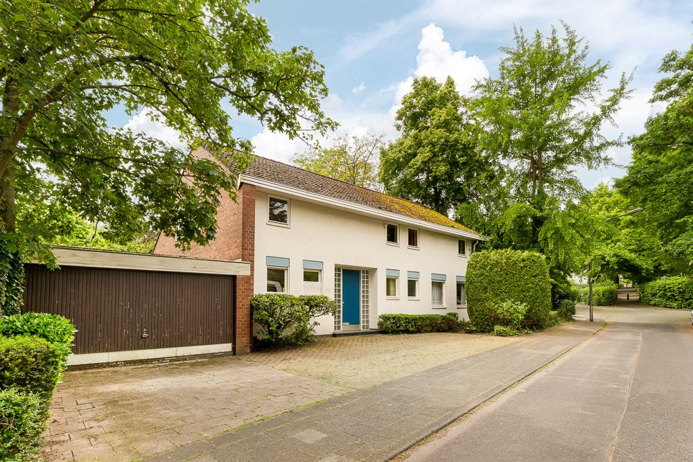 Zweistöckiges Wohnhaus mit Garage, umgeben von Bäumen und Hecken an einer ruhigen Straße.