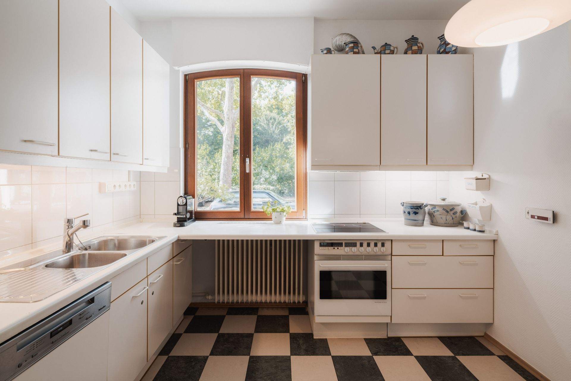 Helle Küche mit weißen Schränken, großem Fenster und schwarz-weiß kariertem Boden.