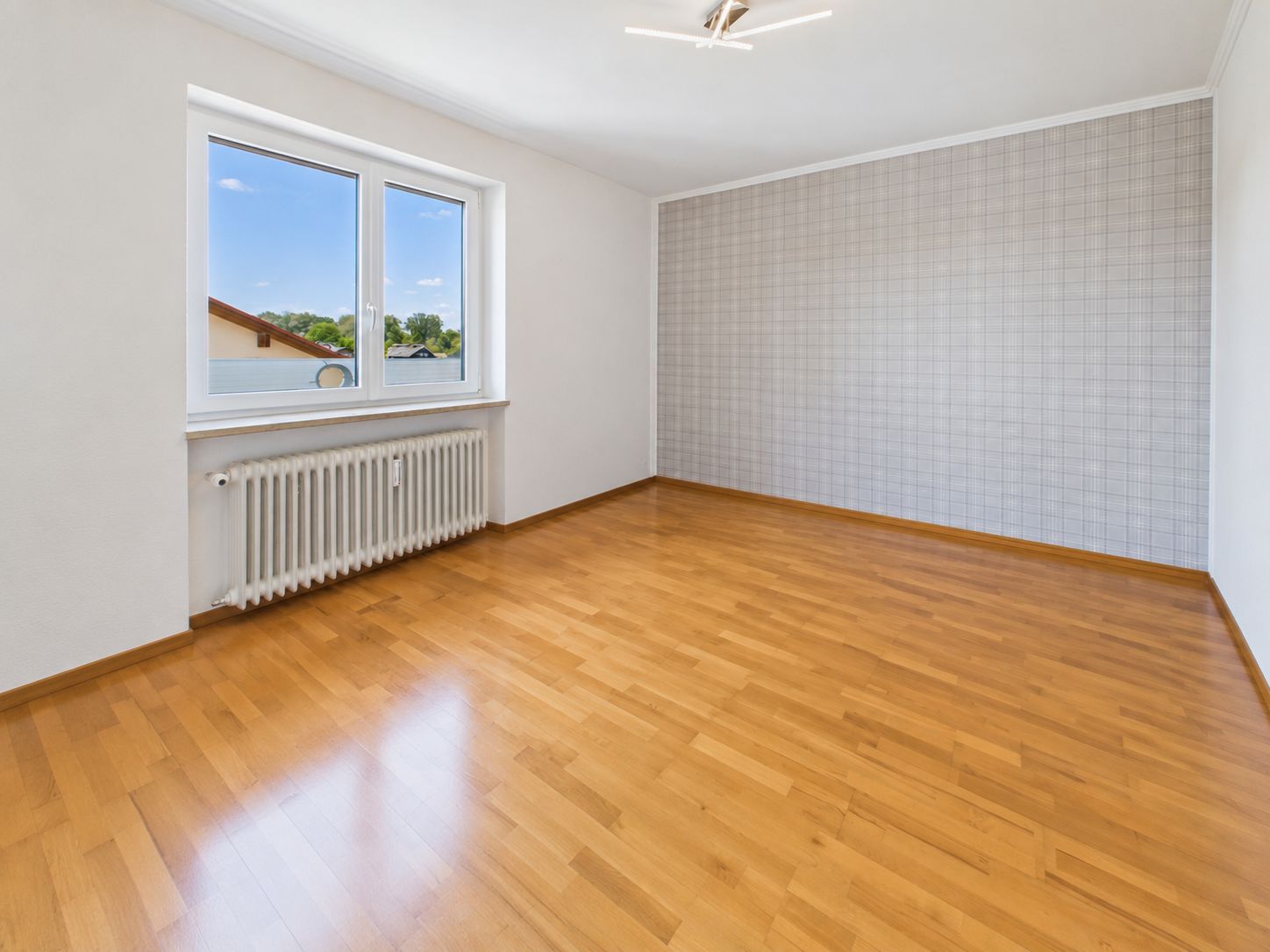 Leeres Zimmer mit Holzfußboden, großem Fenster und gemusterter Tapete an einer Wand.