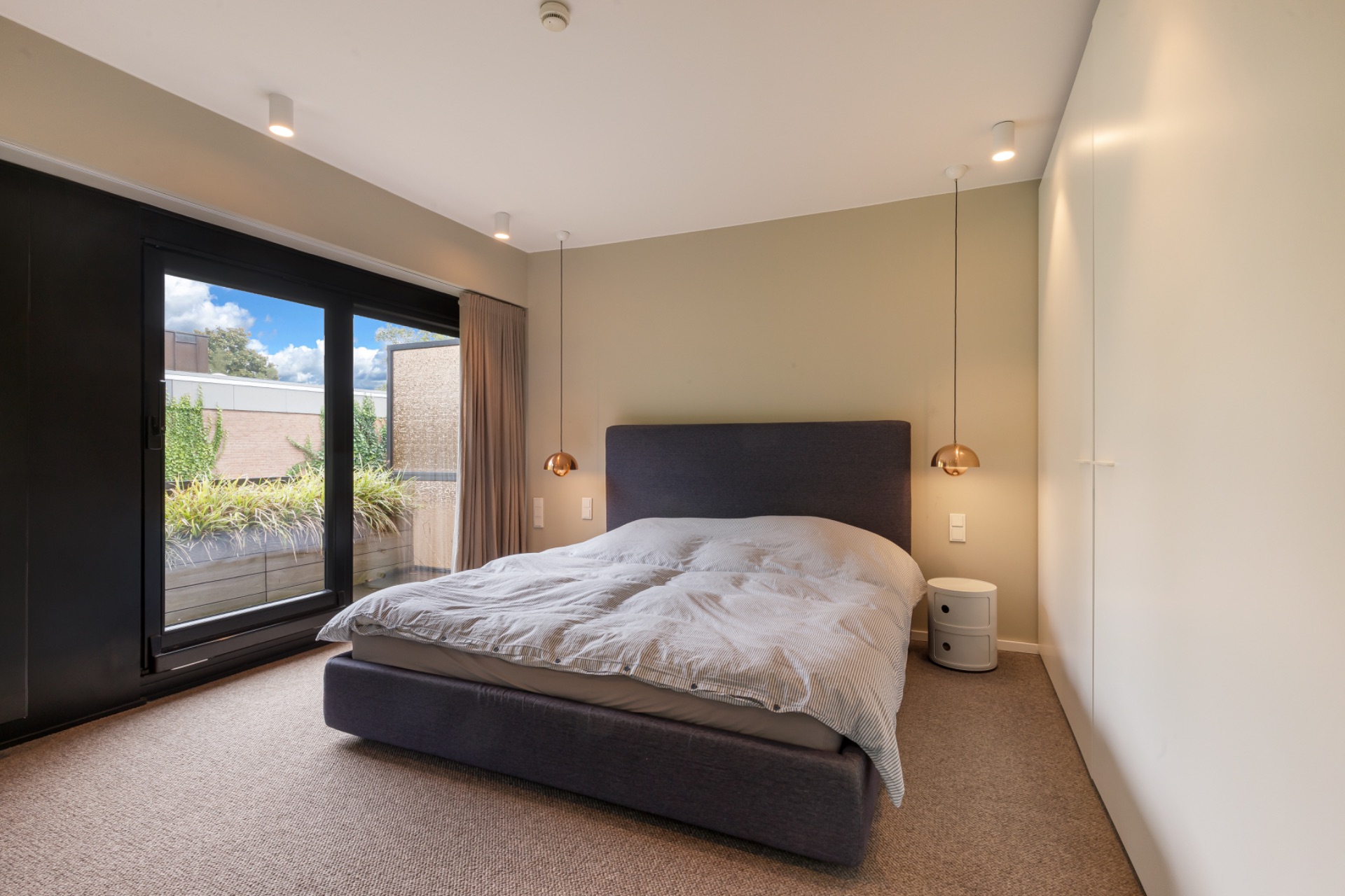 Modernes Schlafzimmer mit Doppelbett, Nachttisch und Zugang zu einem Balkon mit Pflanzen.