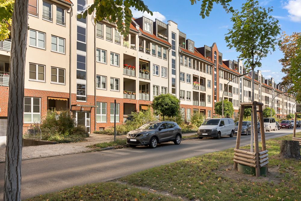 Mehrstöckiges Wohnhaus mit Balkonen, parkenden Autos und Bäumen an einer ruhigen Straße.