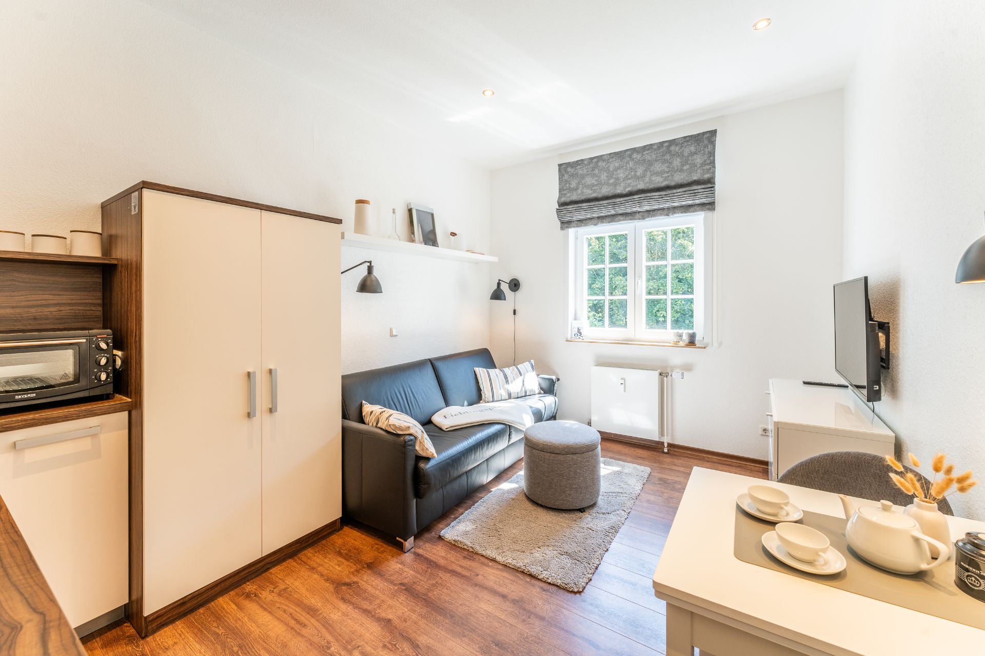Helles Wohnzimmer mit Sofa, Teppich, Ottomane, TV, Esstisch und großem Fenster.
