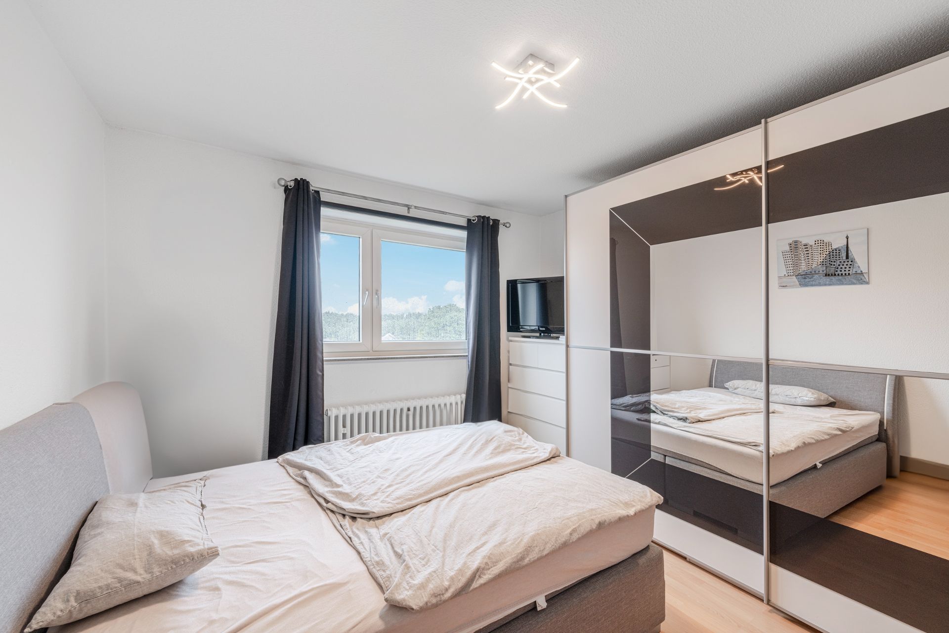 Helles Schlafzimmer mit großem Bett, Kleiderschrank mit Spiegel und Fenster mit Vorhängen.