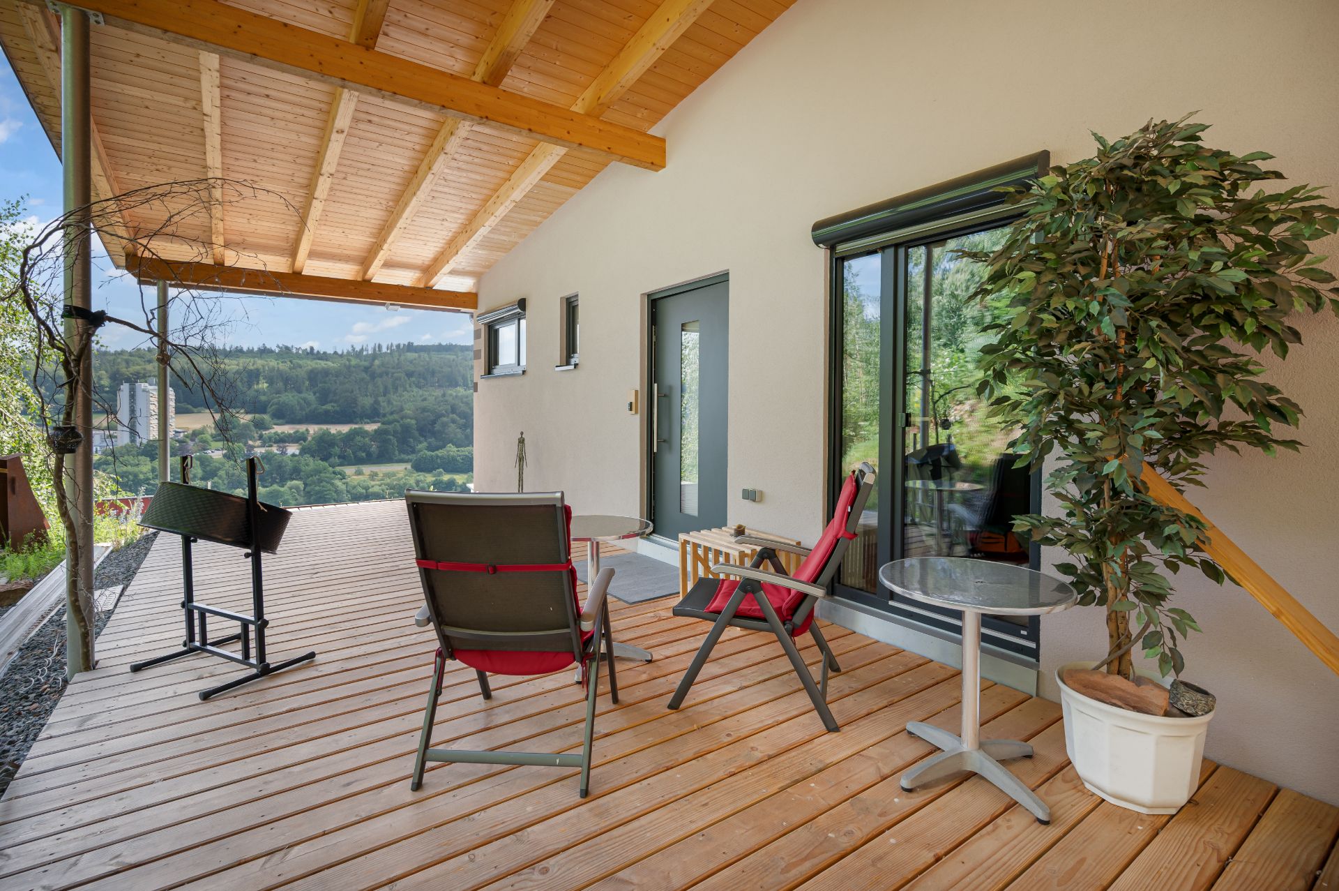 Überdachte Holzterrasse mit zwei Stühlen, kleinem Tisch, Grill und Topfpflanze vor einem Haus.