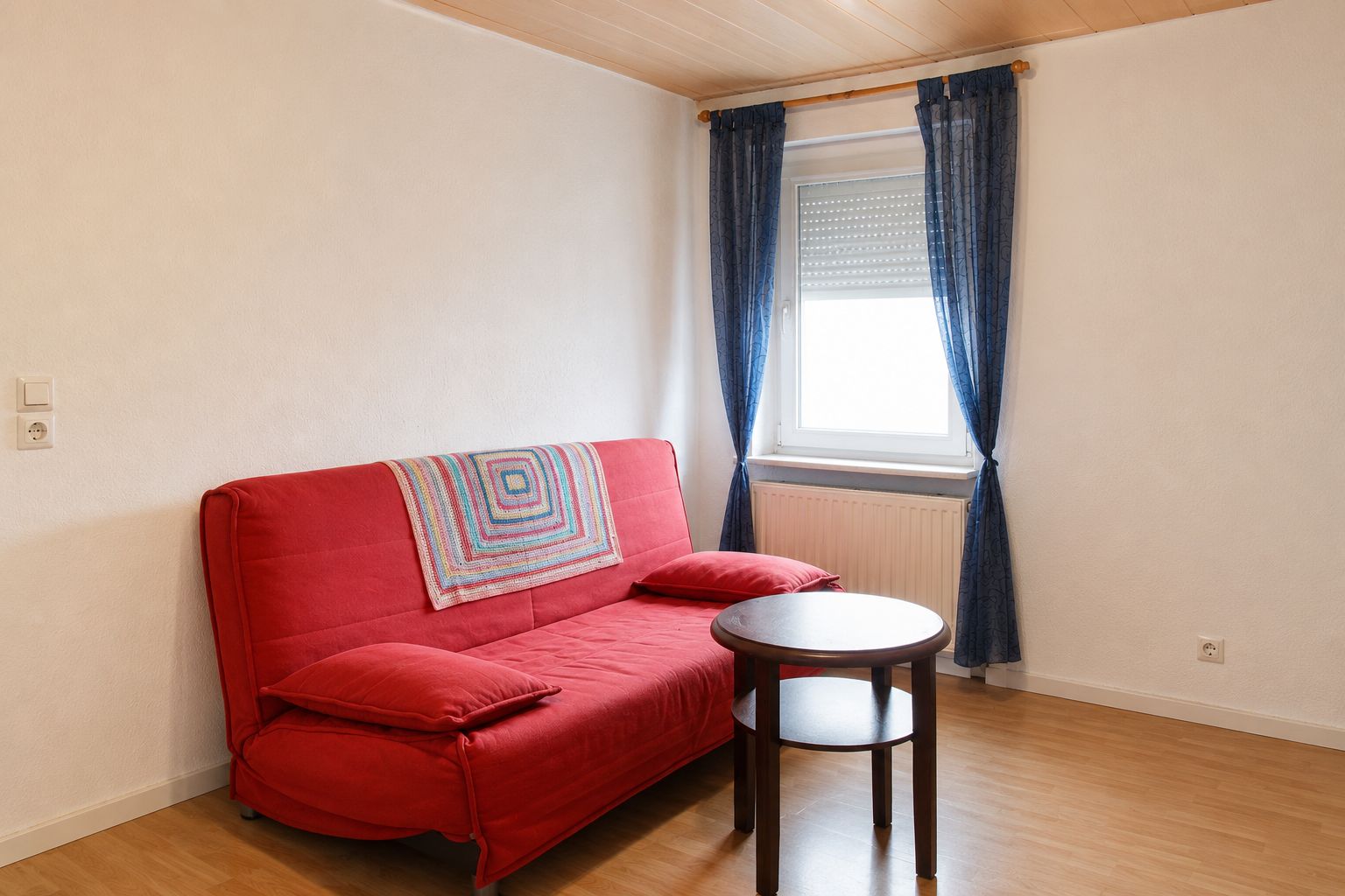 Wohnzimmer mit rotem Sofa, rundem Holztisch und Fenster mit blauen Vorhängen.