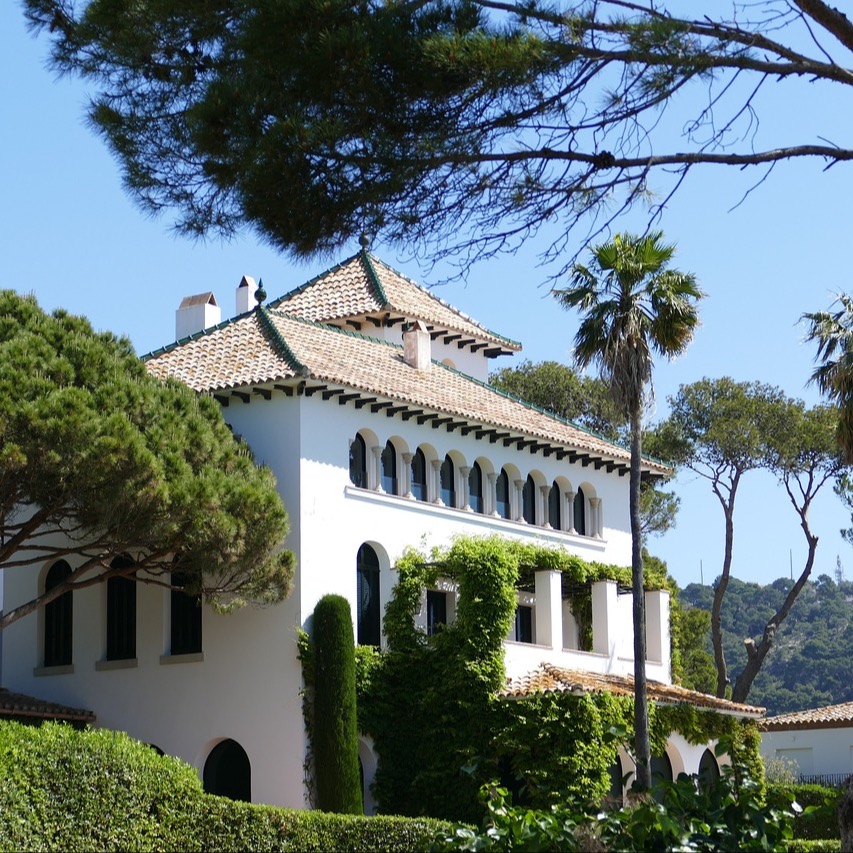 Weißes Wohnhaus mit Ziegeldach, Rundbogenfenstern und viel Grünbewuchs, umgeben von Bäumen.