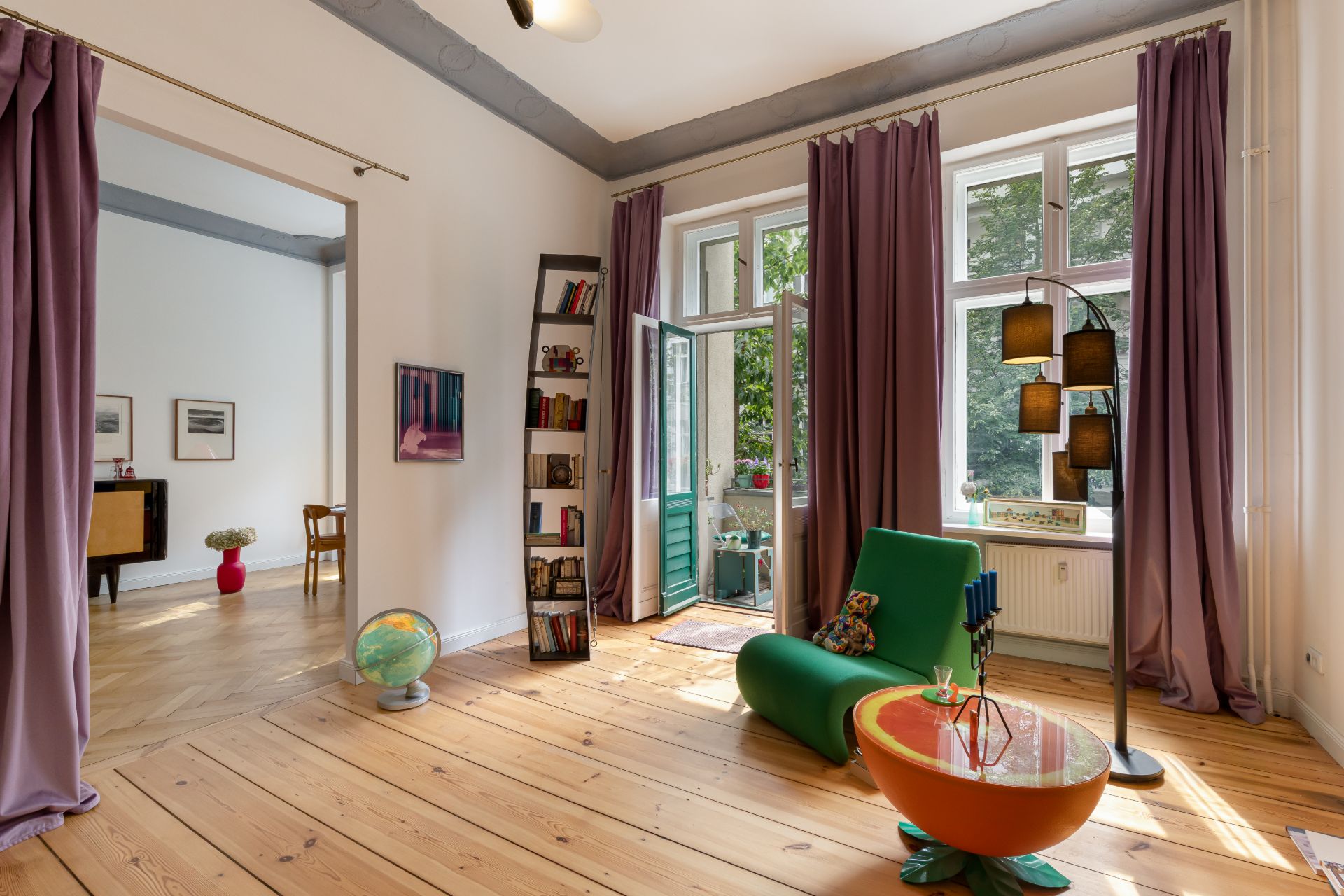 Helles Wohnzimmer mit Holzboden, violetten Vorhängen, grünem Sessel und Globus, große Fenster zum Garten.