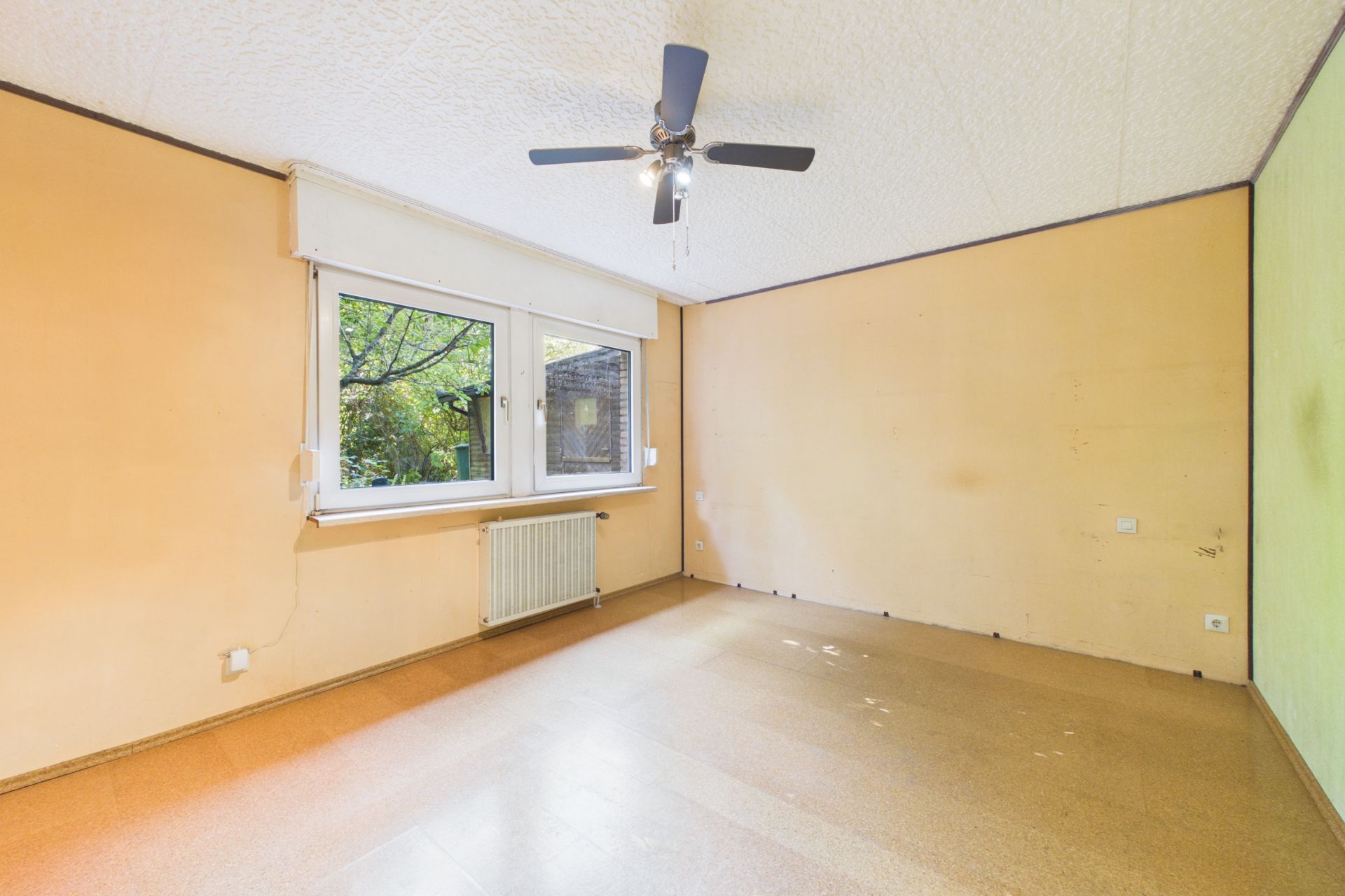 Leerer Raum mit beigem Wänden, großem Fenster, Heizkörper und Deckenventilator.