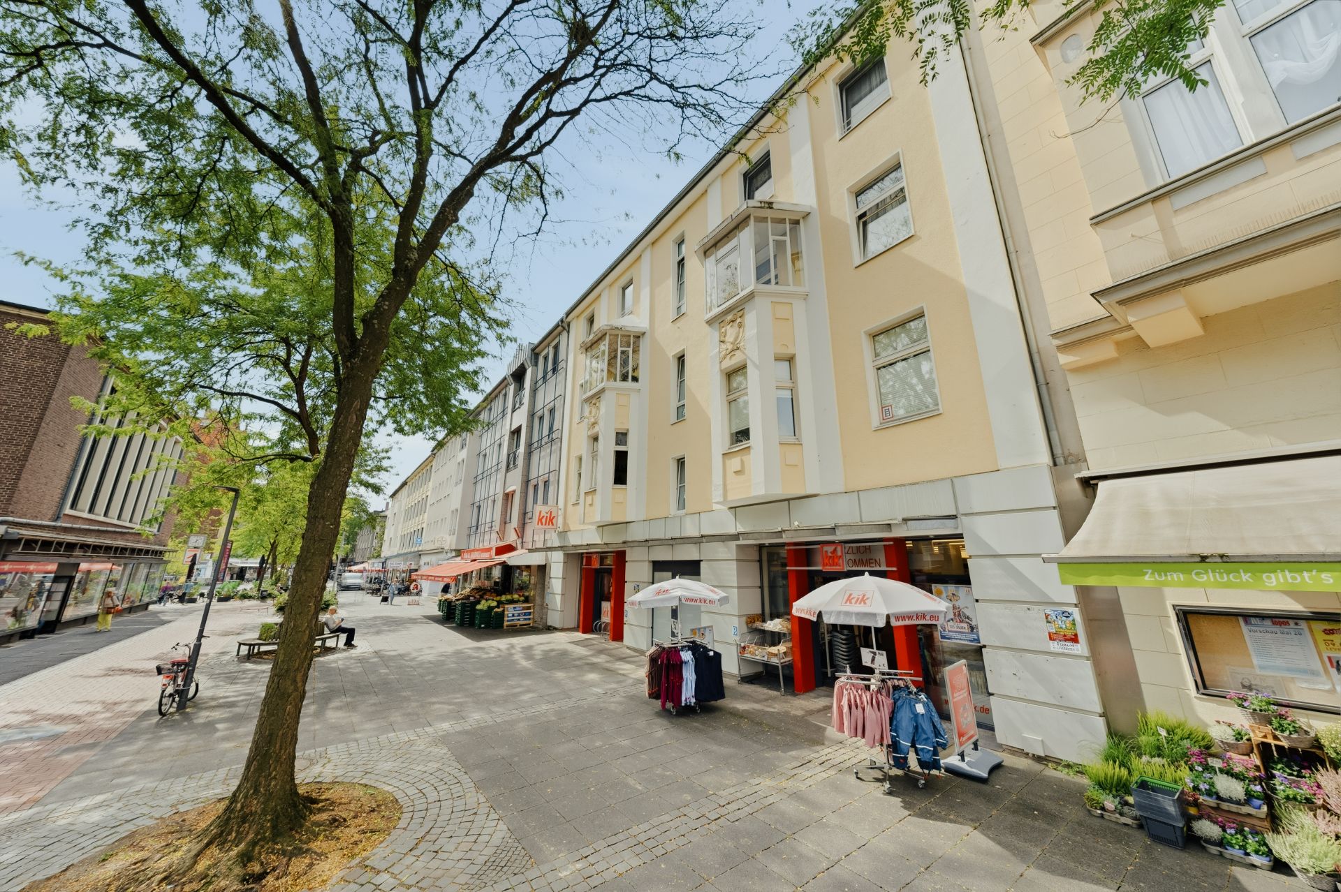 Mehrstöckiges Wohnhaus mit Läden im Erdgeschoss, Baum und Gehweg davor.