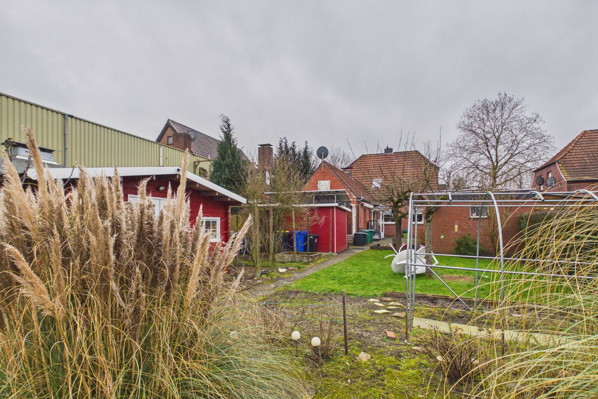 Garten mit Rasen, Ziergräsern, Schaukel und roten Gebäuden im Hintergrund.