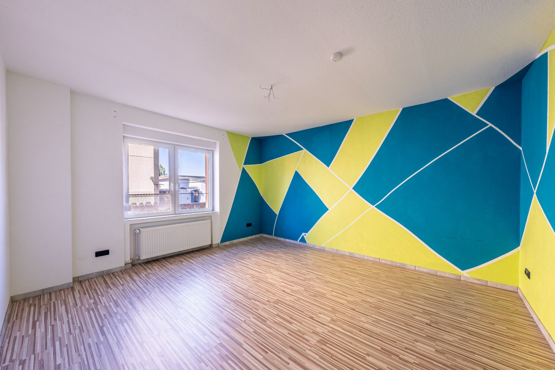 Leerer Raum mit Holzboden, Fenster und bunter geometrischer Wandmalerei in Blau und Gelb.