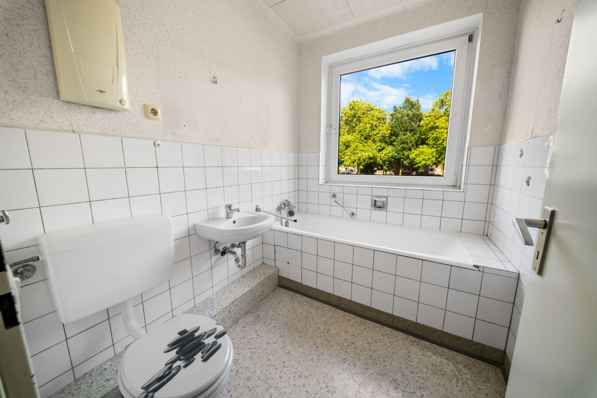 Helles Badezimmer mit Badewanne, Waschbecken, Toilette und Fenster mit Blick ins Grüne.