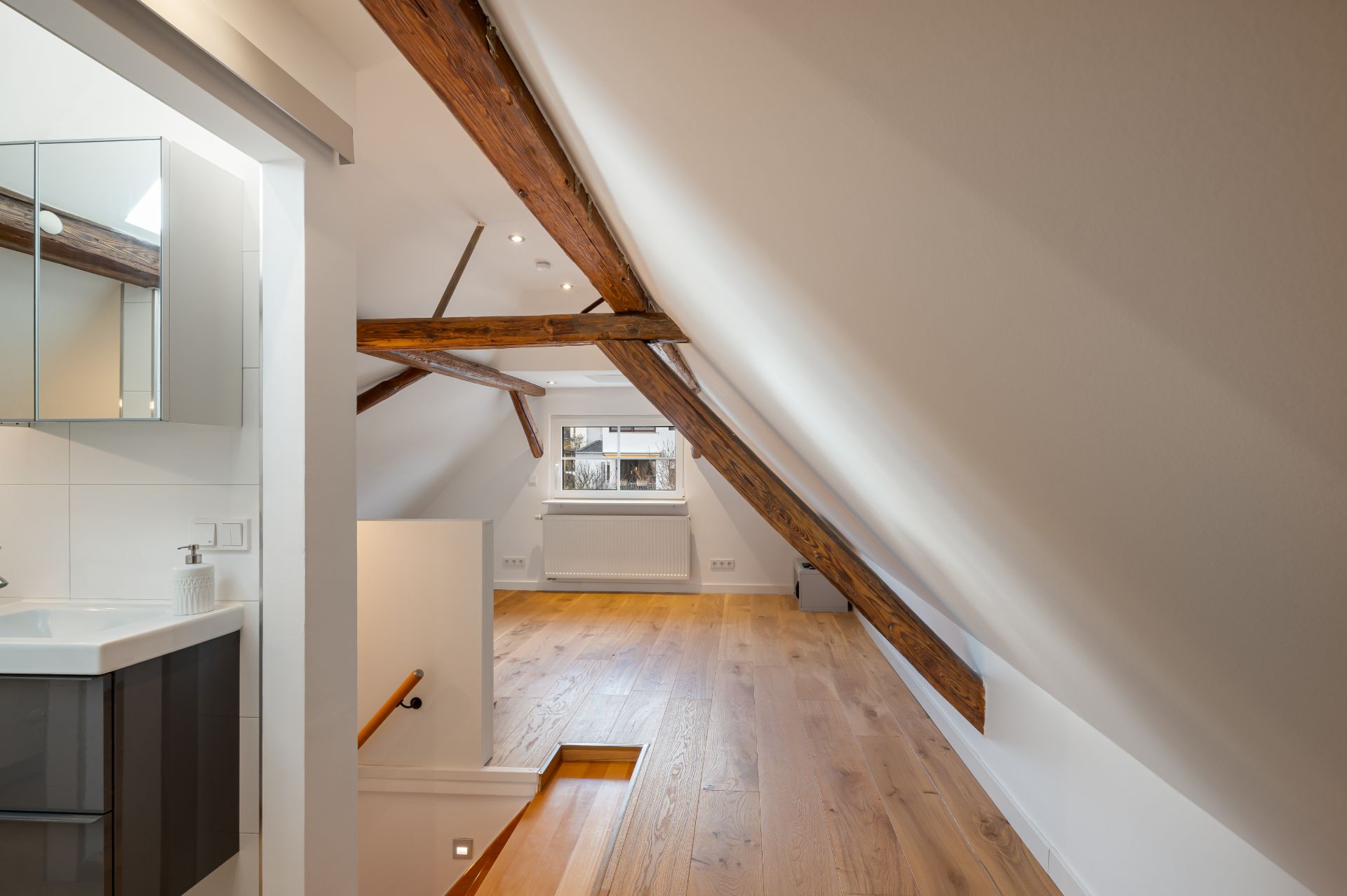 Dachgeschossraum mit sichtbaren Holzbalken, Holzfußboden, Fenster und kleinem Waschbecken.