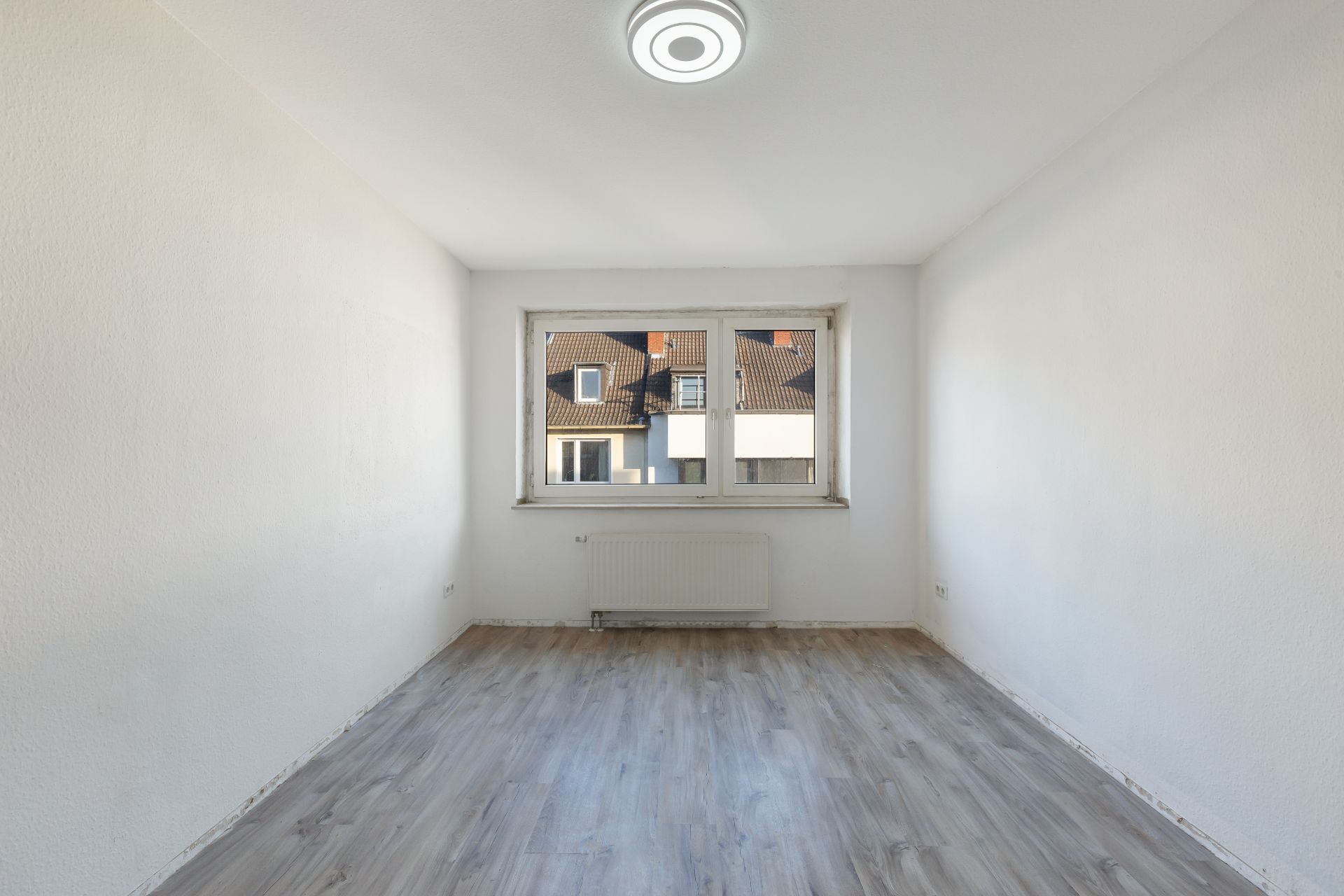Leerer Raum mit hellem Boden, weiß gestrichenen Wänden und Fenster mit Blick auf Nachbarhäuser.