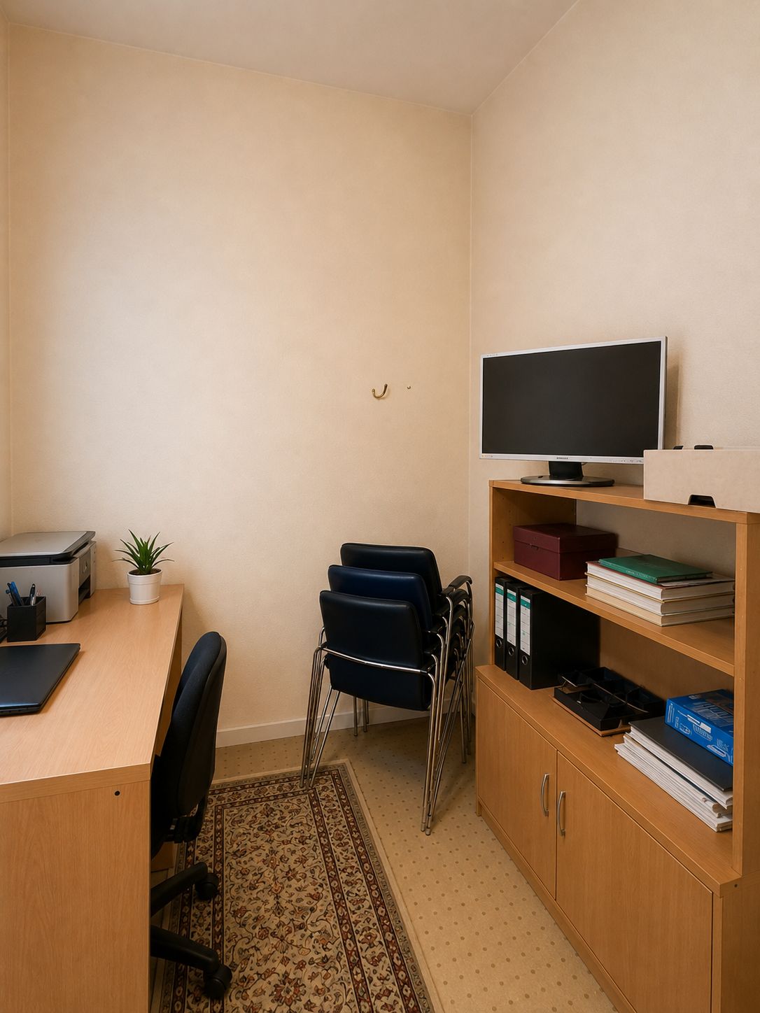 Kleines Arbeitszimmer mit Schreibtisch, Stuhl, Regal, Monitor und mehreren gestapelten Stühlen.
