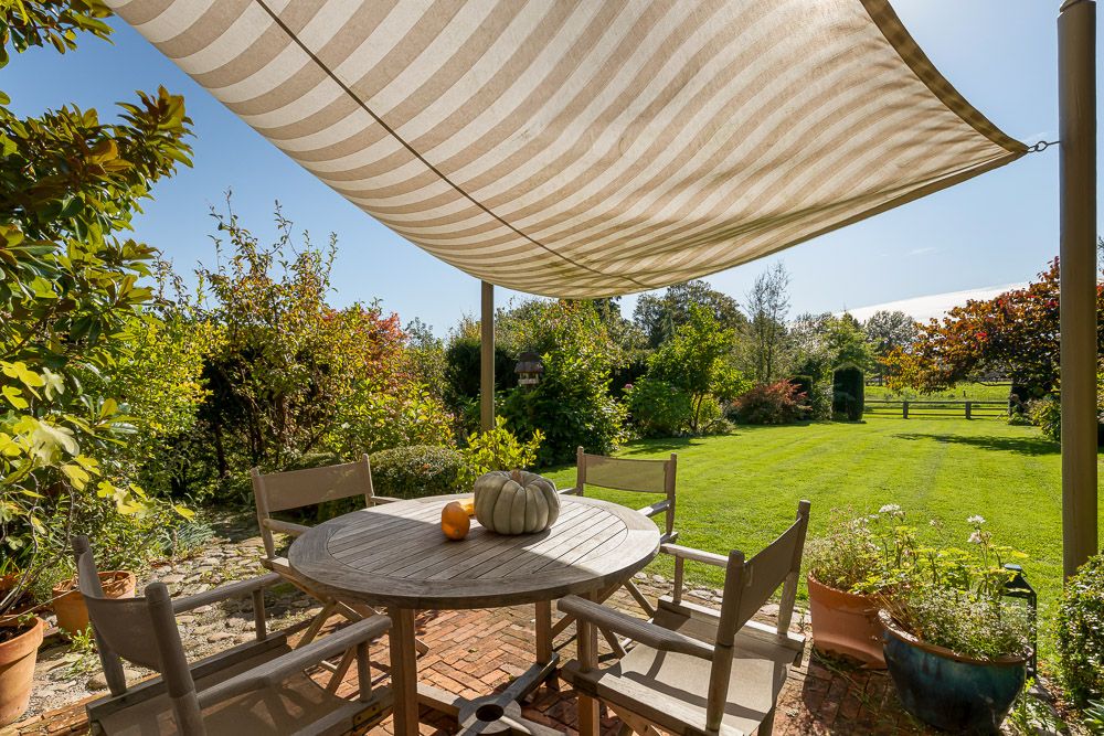 Holztisch mit fünf Stühlen auf Terrasse, Markise darüber, grüner Garten im Hintergrund.